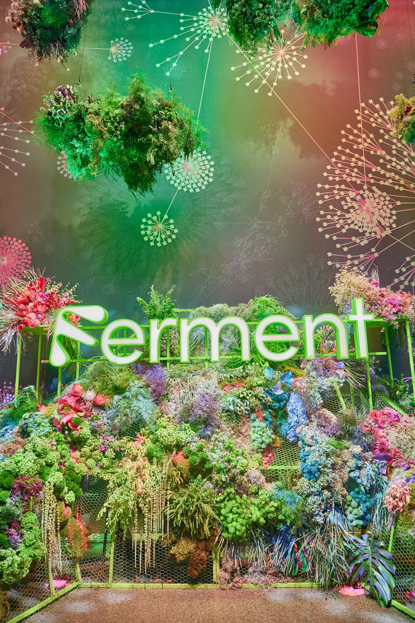 A colorful floral display with the word 'Serment' illuminated in green and white neon lights, set against a vibrant backdrop of various flowers and greenery.