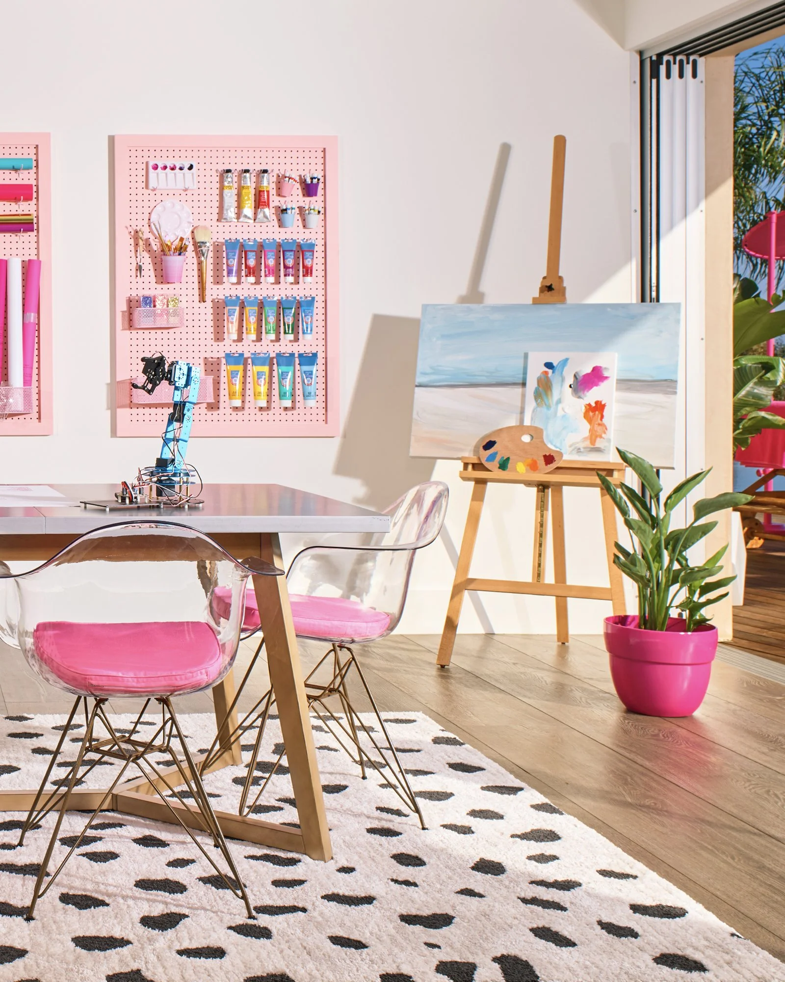 Art studio with a small painting on an easel, paint palettes, brushes, and colored paint tubes on pink pegboards, pink and clear chairs, a patterned rug, and a potted plant near a window with green foliage outside.