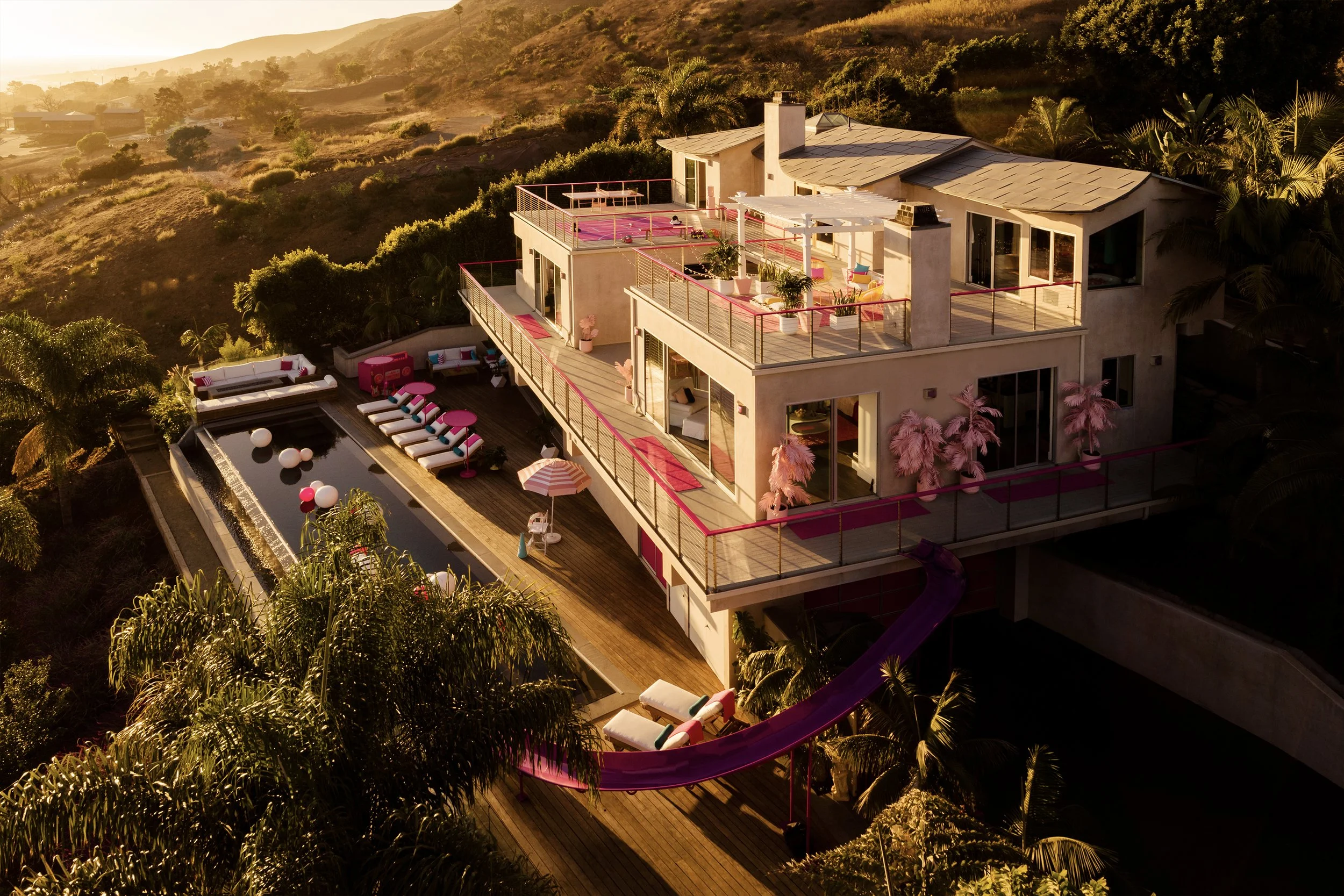 A large house on a hillside with multiple levels and outdoor terraces, decorated with pink accents. There is a swimming pool with lounge chairs, umbrellas, and colorful seating areas. The house is surrounded by lush greenery and palm trees, with a sunset casting a warm glow over the scene.