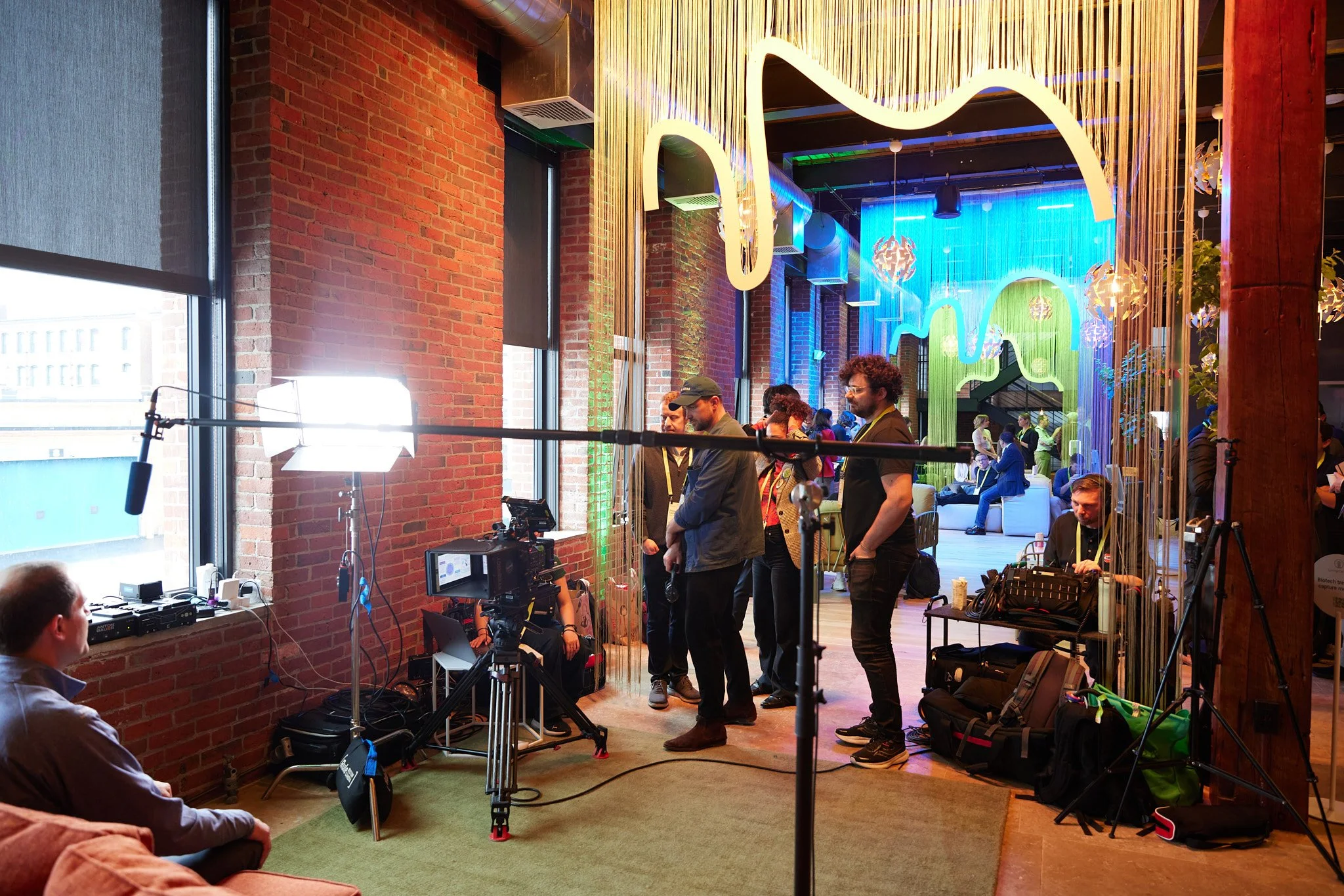 Interior of a modern industrial-style event space with brick walls, large windows with roller blinds, colorful hanging lights, and people setting up or preparing for an event, including cameras and electronic equipment.