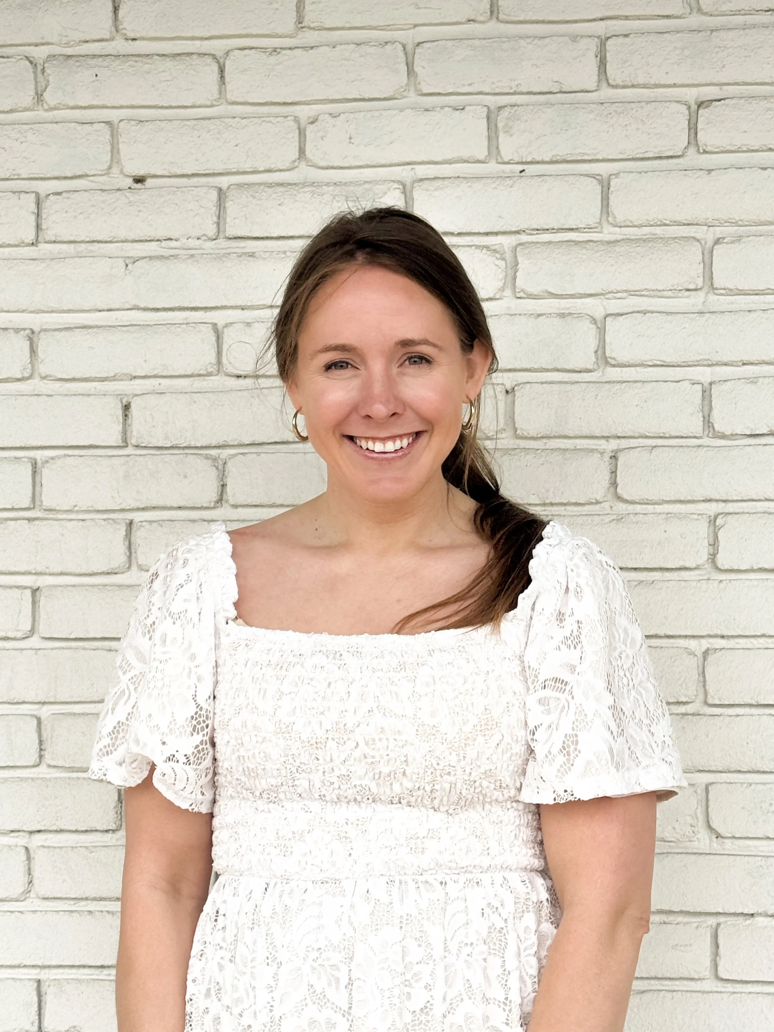A woman with light skin, brown hair in a loose side braid, and hoop earrings smiling, wearing a white lace dress with puffed sleeves, standing in front of a white brick wall.