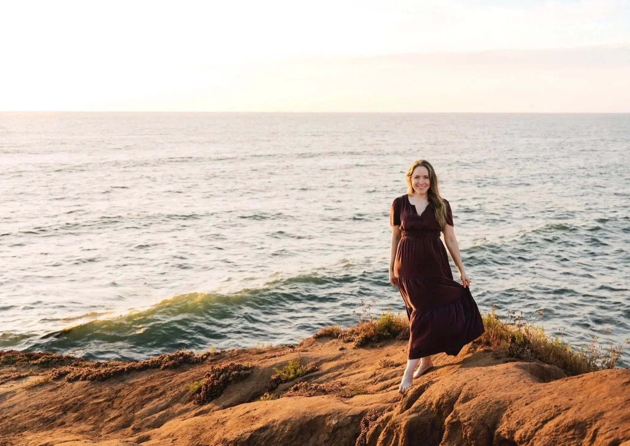 A woman in a brown dress standing on a rocky shoreline, smiling, with ocean waves and a bright sky in the background.