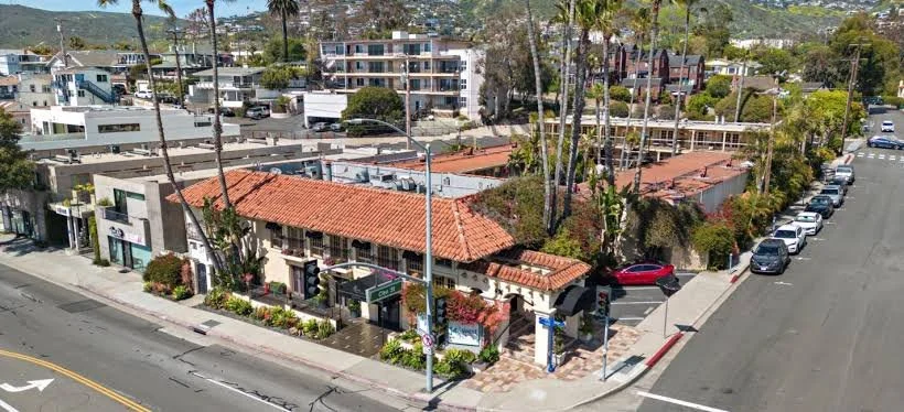 Aerial view of a city street with shops, trees, parked cars, and multi-story residential buildings in the background.