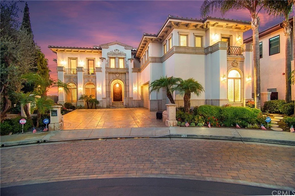 Luxurious two-story mansion with bright exterior lighting, palm trees, landscaped garden, and a paved driveway at sunset.