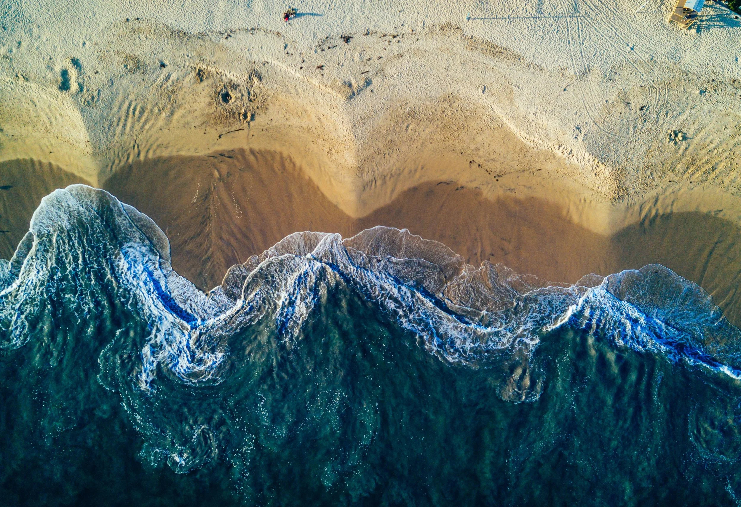 An aerial view of the beach showing waves crashing onto the shore and footprints in the sand, with a few people and umbrellas.