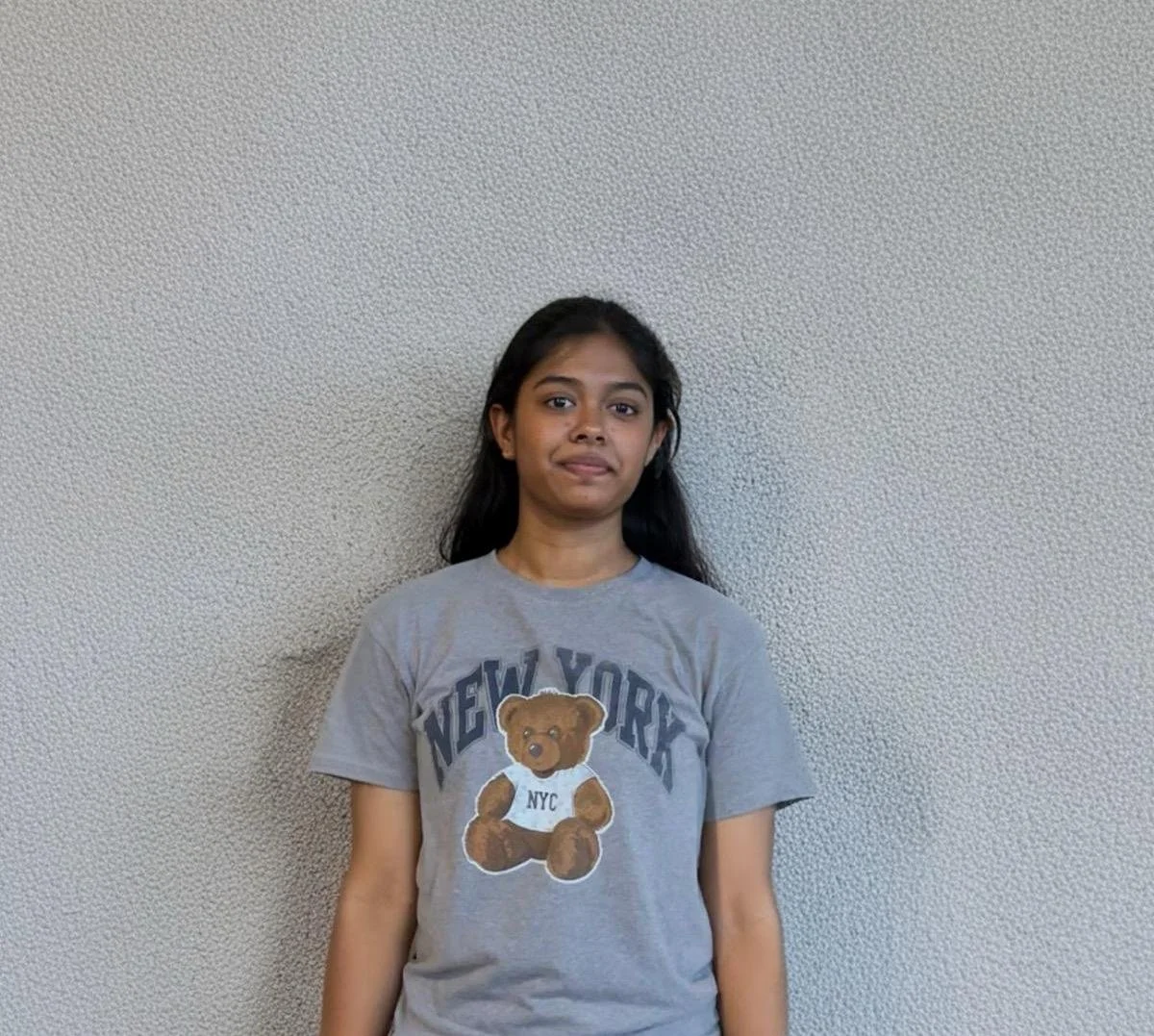 A young girl with long dark hair stands against a plain, textured wall. She is wearing a gray T-shirt with a bear graphic and the words "New York" and "NYC" on it.