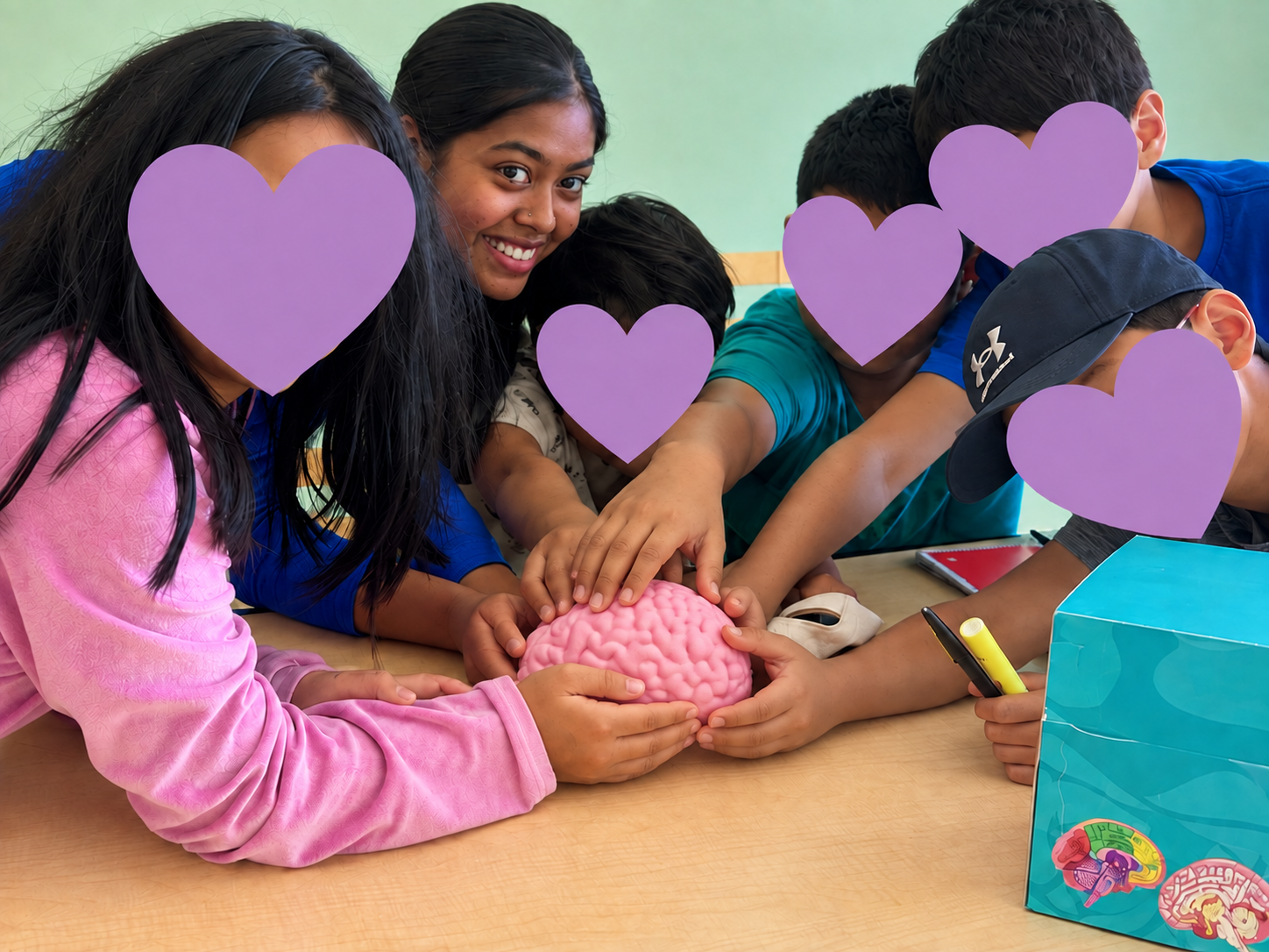 Dhiya and workshop participants holding a pink foam brain model at Cambrian Library; children’s faces covered with purple hearts.