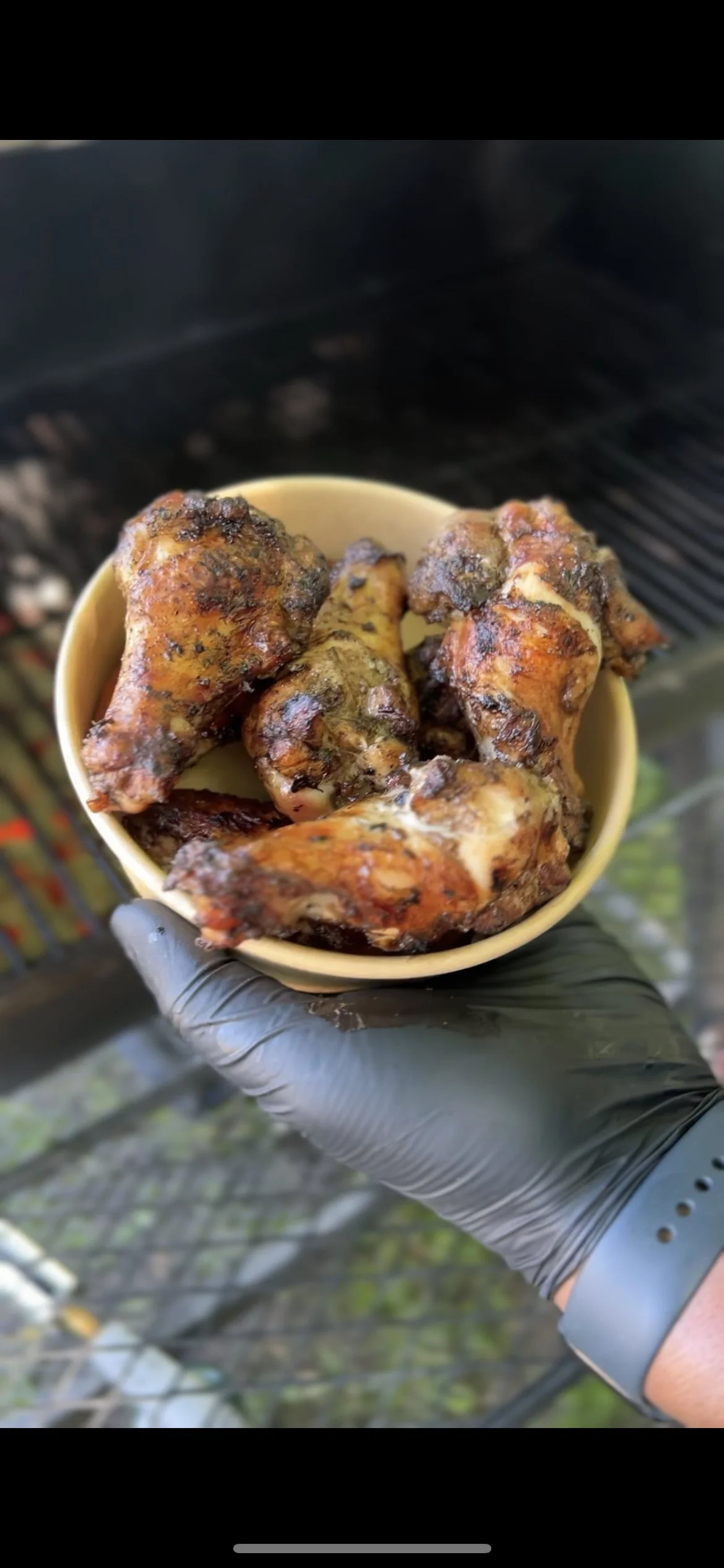 A hand wearing a black glove holding a yellow bowl with grilled chicken drumsticks, outdoors.