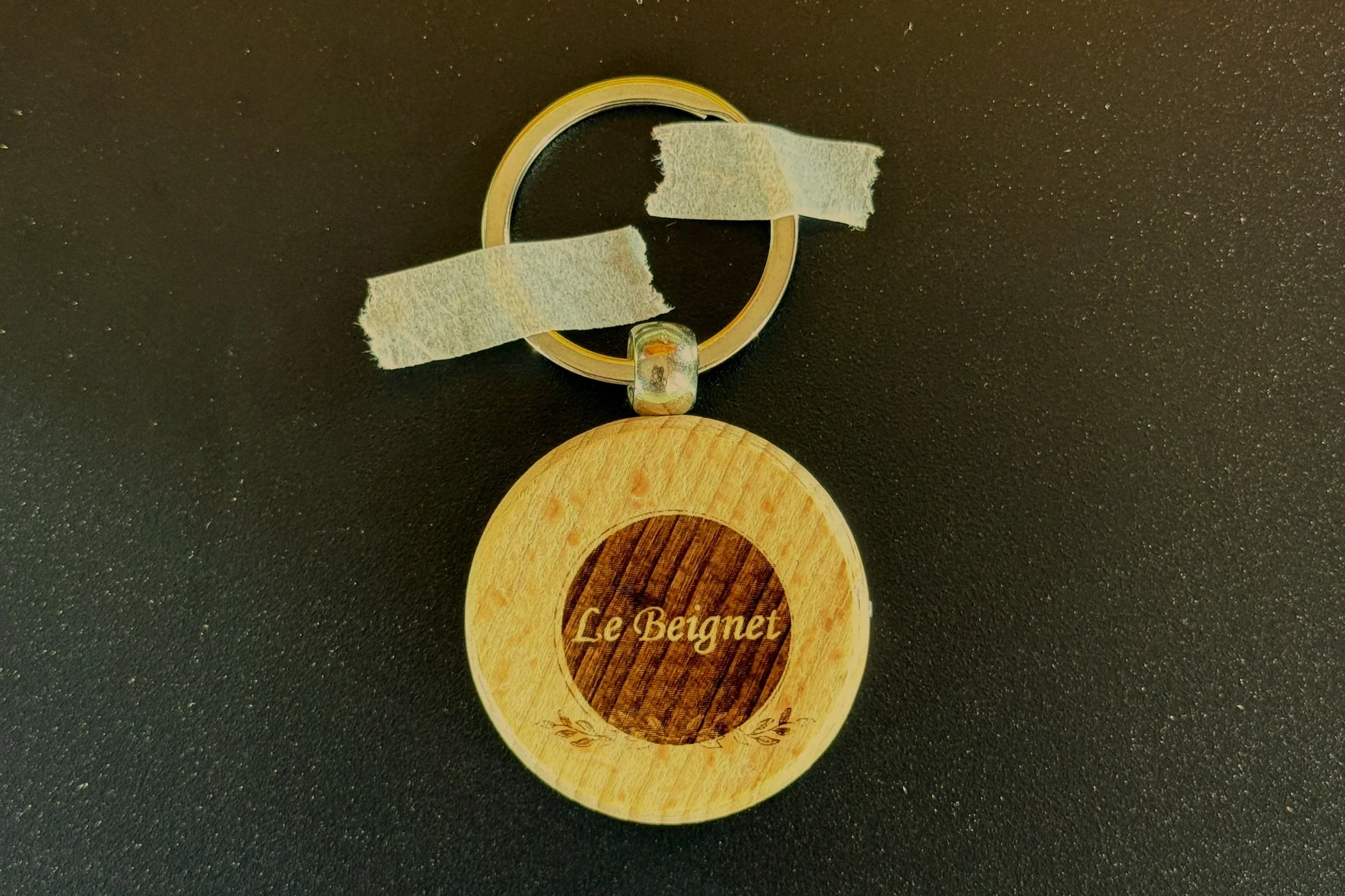A closed small tin can with a pull-tab lid placed on a black surface, with white tape on the tab. Below the can, there is a round wooden coaster with a logo that reads 'Le Beignet' and an illustration of a beignet.