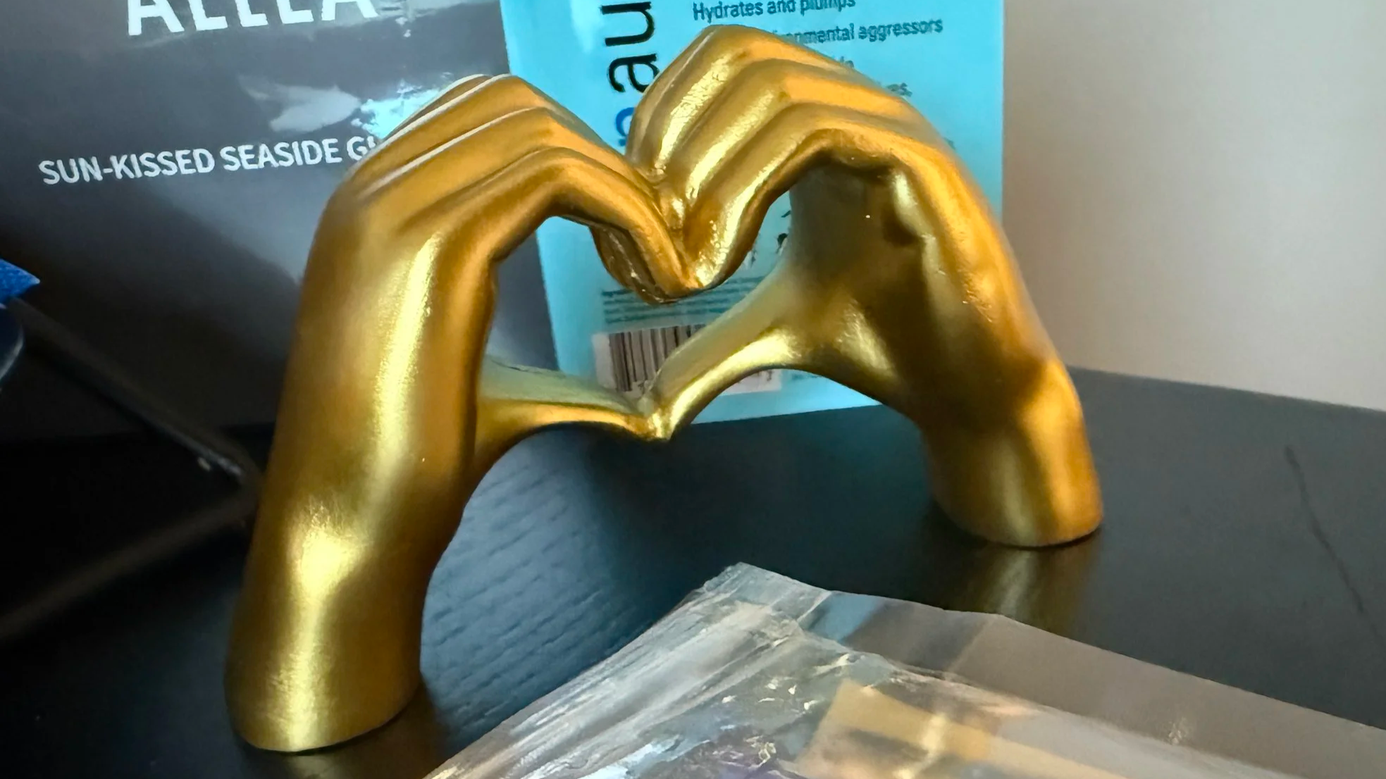 A gold-colored hand sculpture making a heart shape with its fingers, placed on a black surface with some packaging or wrappers and a blue box in the background.