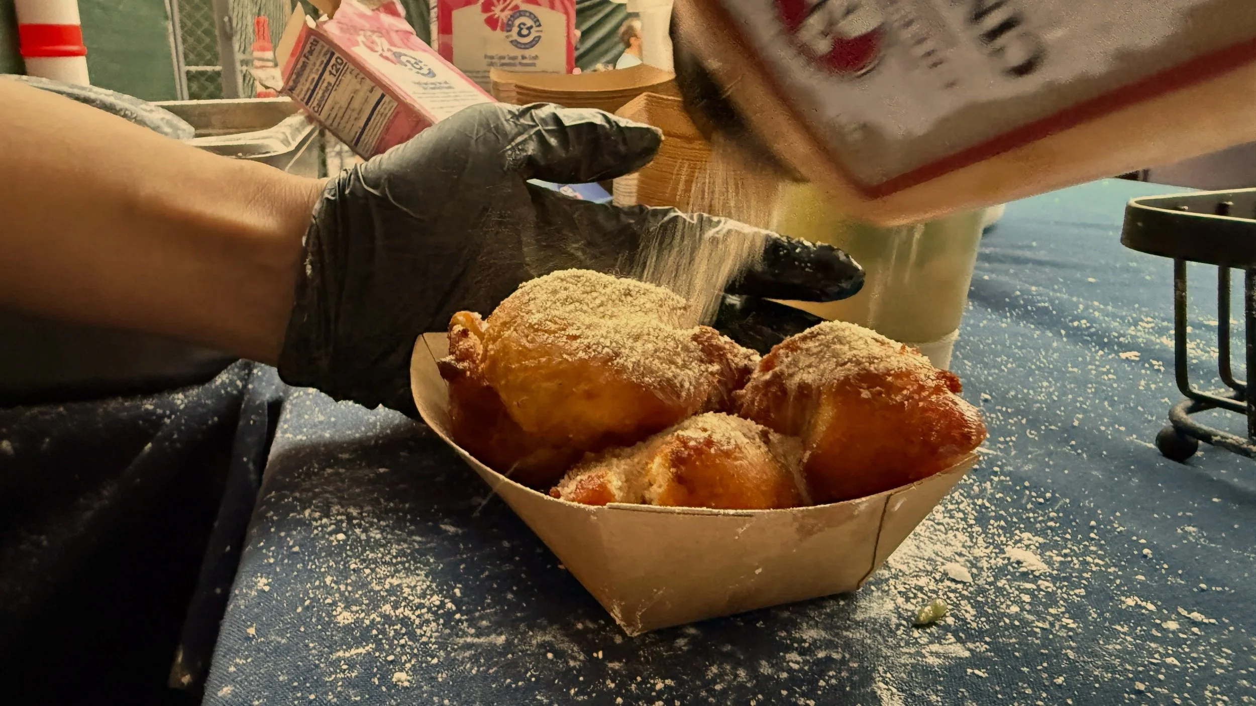 A person wearing a black glove holding a paper container of fried food topped with powdered sugar, with a table and outdoor setting in the background.