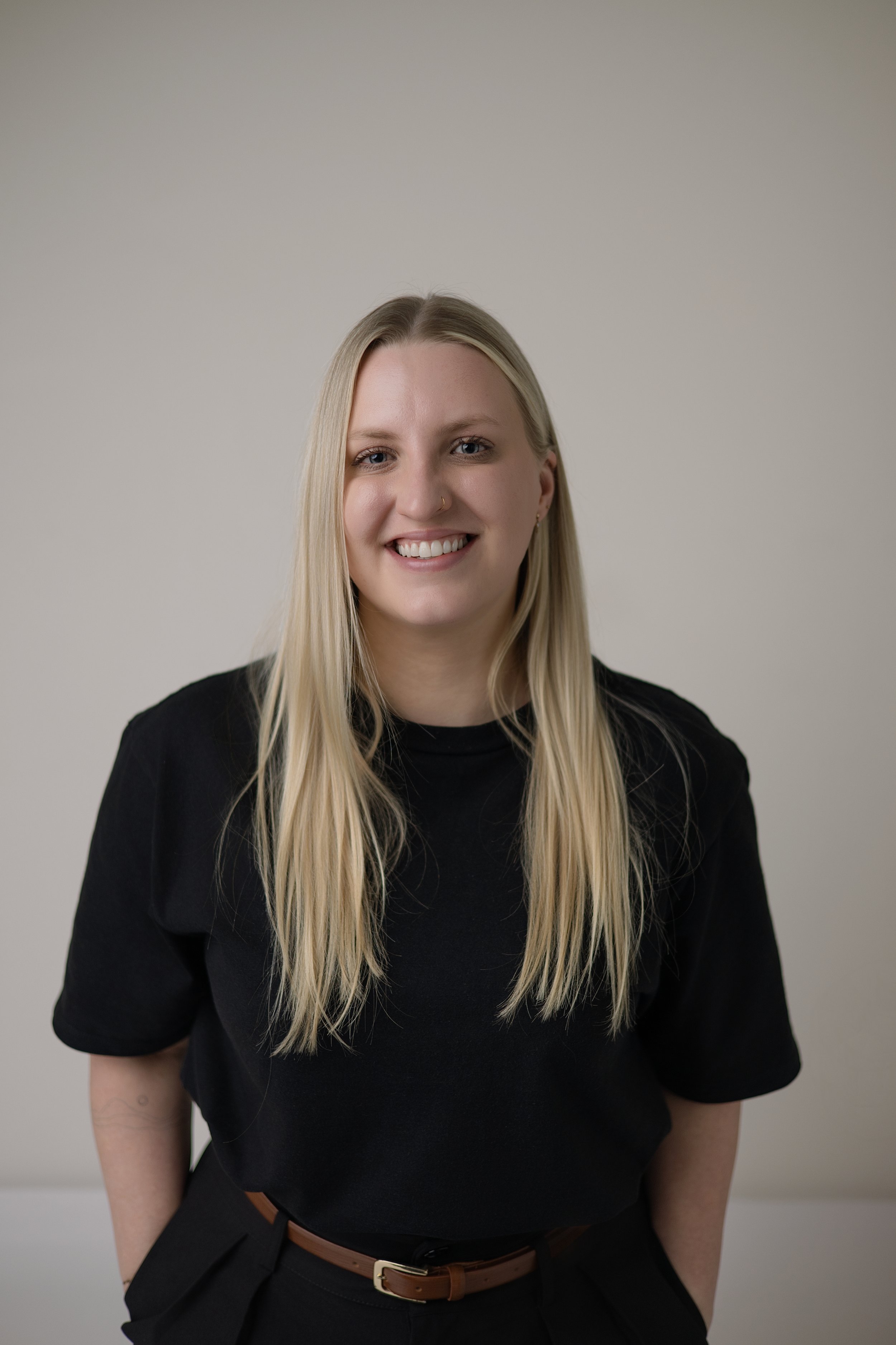 Jen Taylor Psychologist Brisbane EMDR PTSD anxiety A young woman with long blonde hair, smiling, wearing a black t-shirt and black pants with a brown belt, standing in front of a plain light-colored background.
