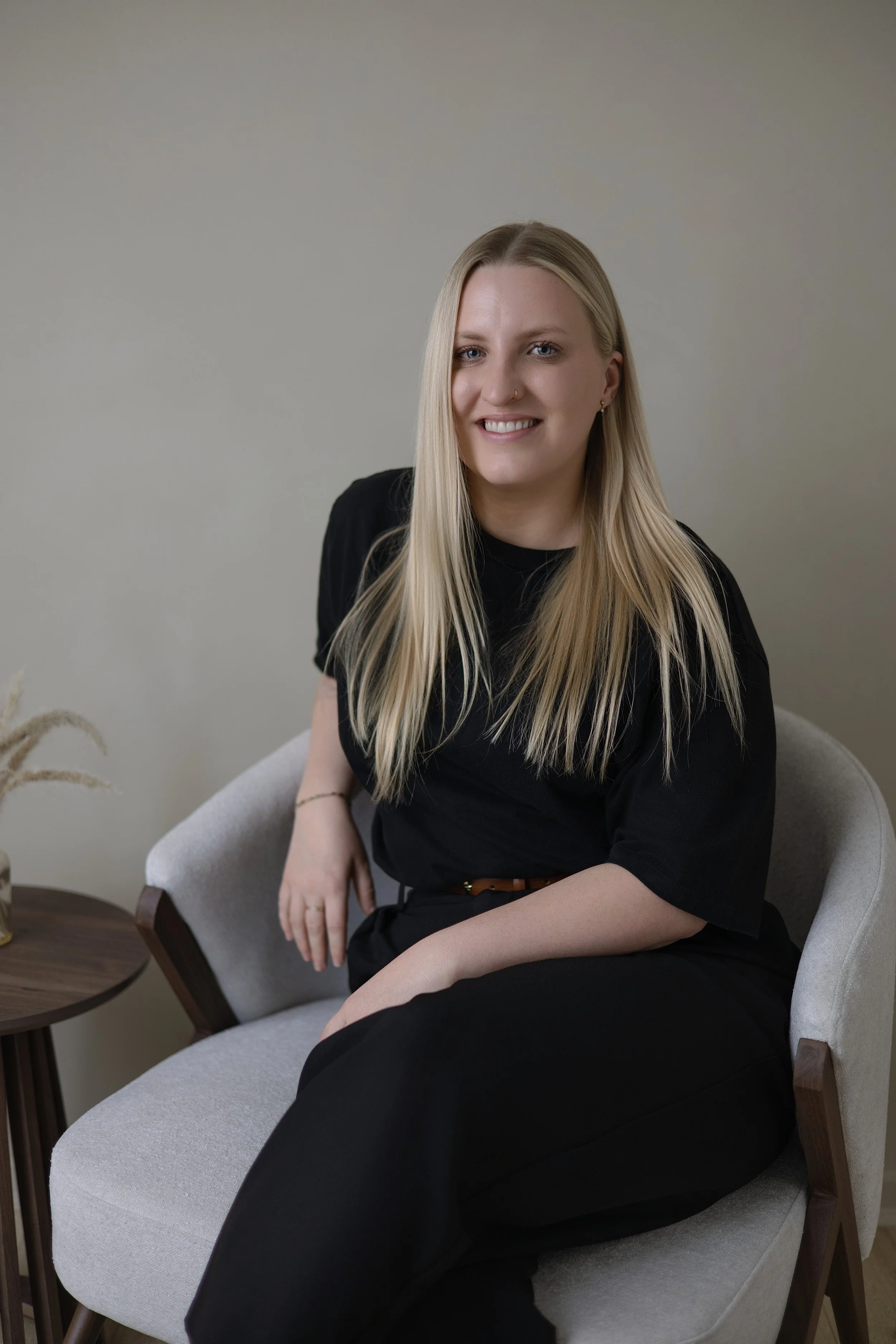 Jen Taylor Psychologist Brisbane EMDR PTSD anxiety A smiling woman with long blonde hair seated in a light gray armchair next to a small round wooden side table with a gold vase containing dried plants, against a plain beige wall.