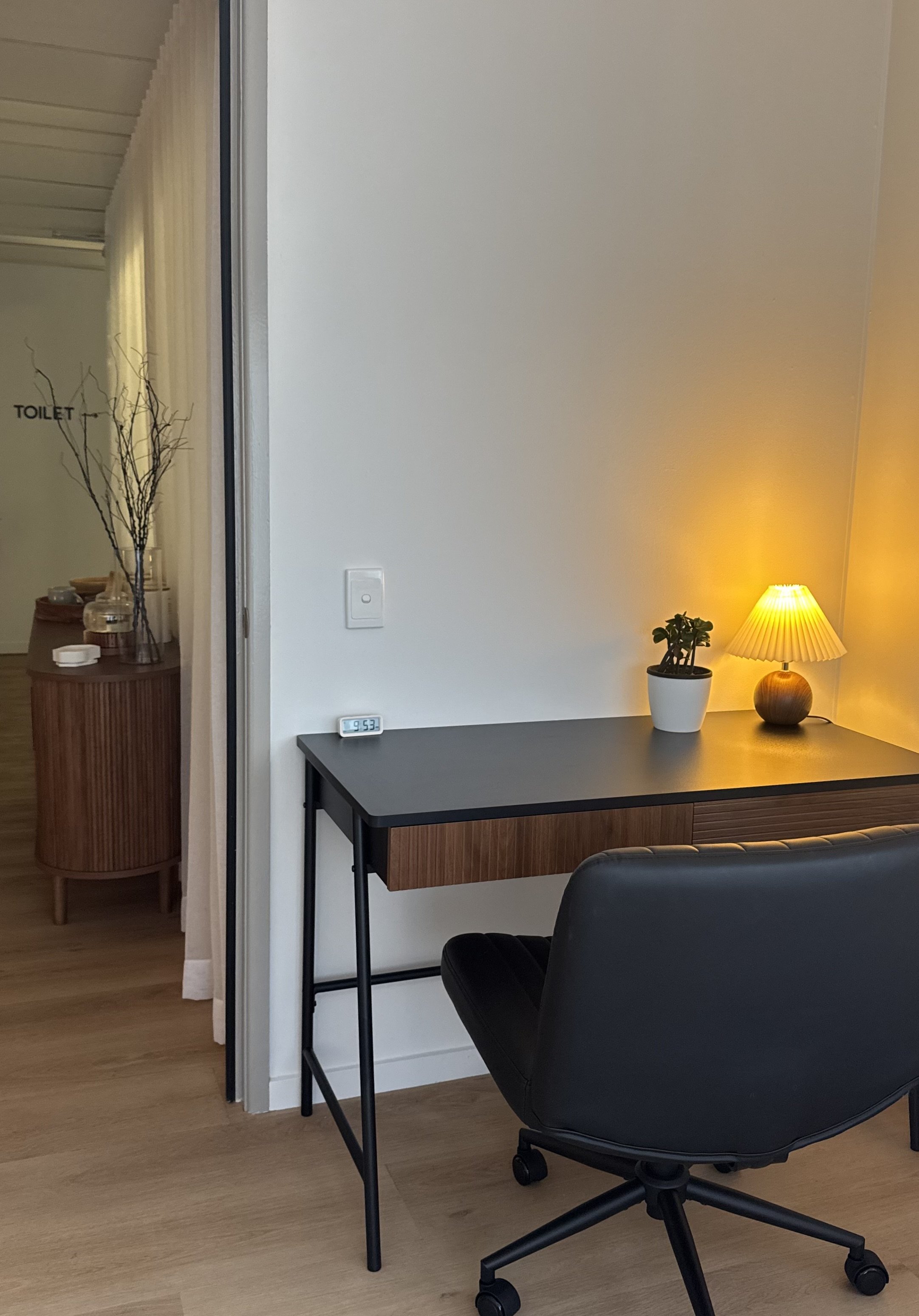Jen Taylor Psychologist Brisbane EMDR PTSD anxiety A room corner with a black desk, a potted plant, a lamp with a yellow shade, a digital clock showing 9:53, and a black office chair.