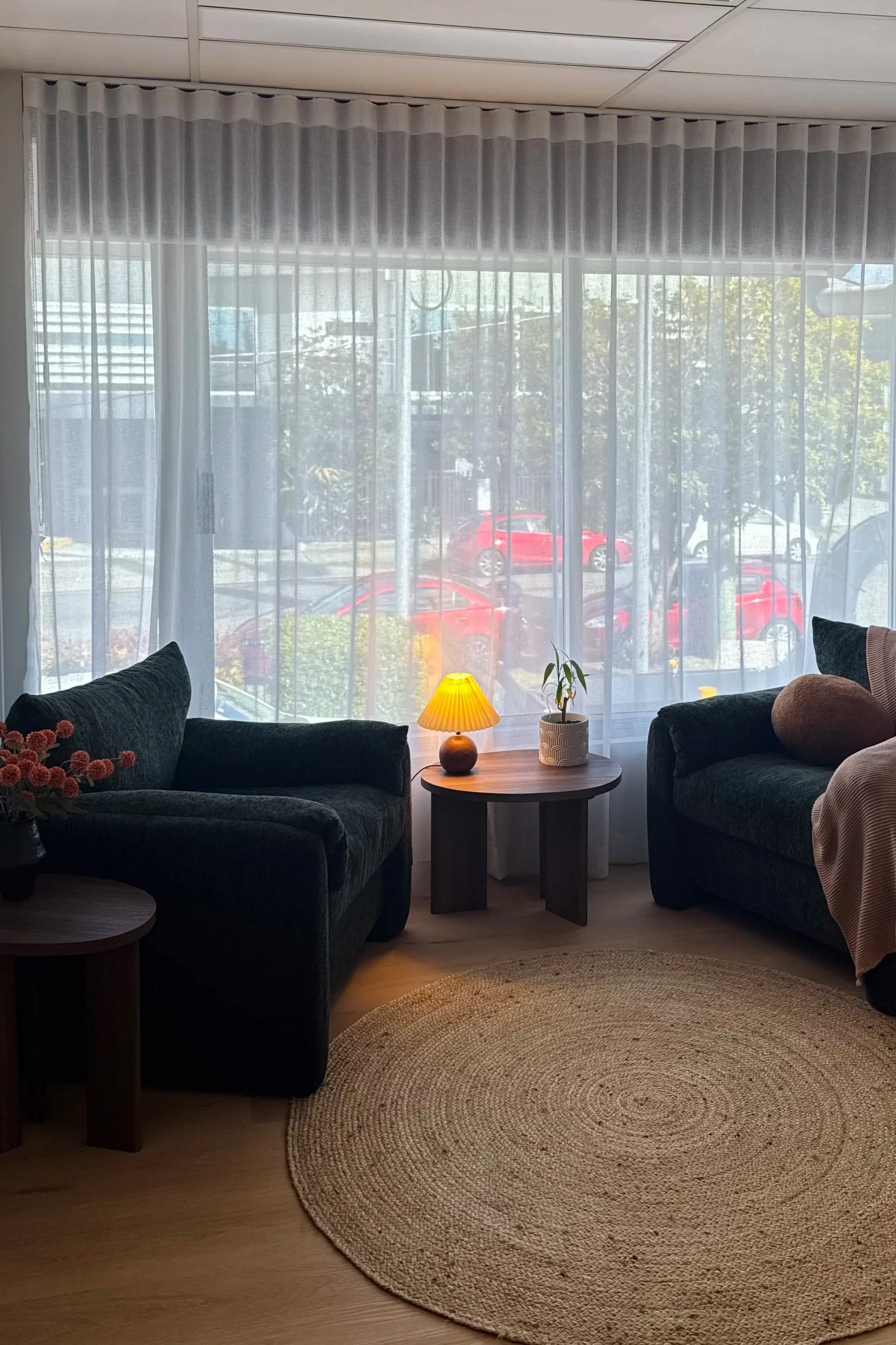 Jen Taylor Psychologist Brisbane EMDR PTSD anxiety Living room with two dark green armchairs, two small side tables, a rug, a window with white sheer curtains, a small lamp, and a potted plant.