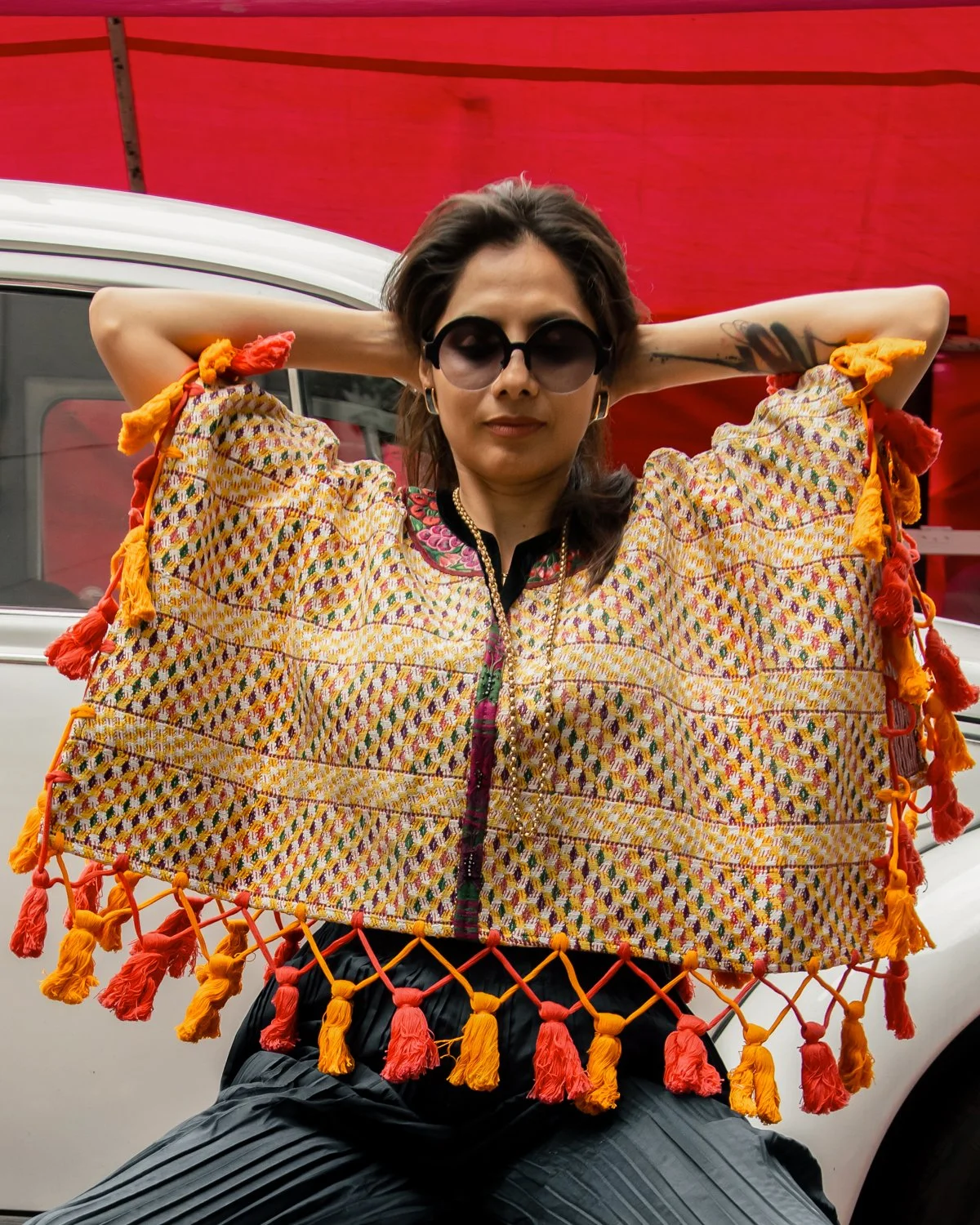 A woman wearing large sunglasses and a colorful, patterned garment with tassels holds a vibrant woven shawl with orange, pink, and yellow tassels in front of her. She is standing outdoors in front of a vehicle with a red canopy overhead.