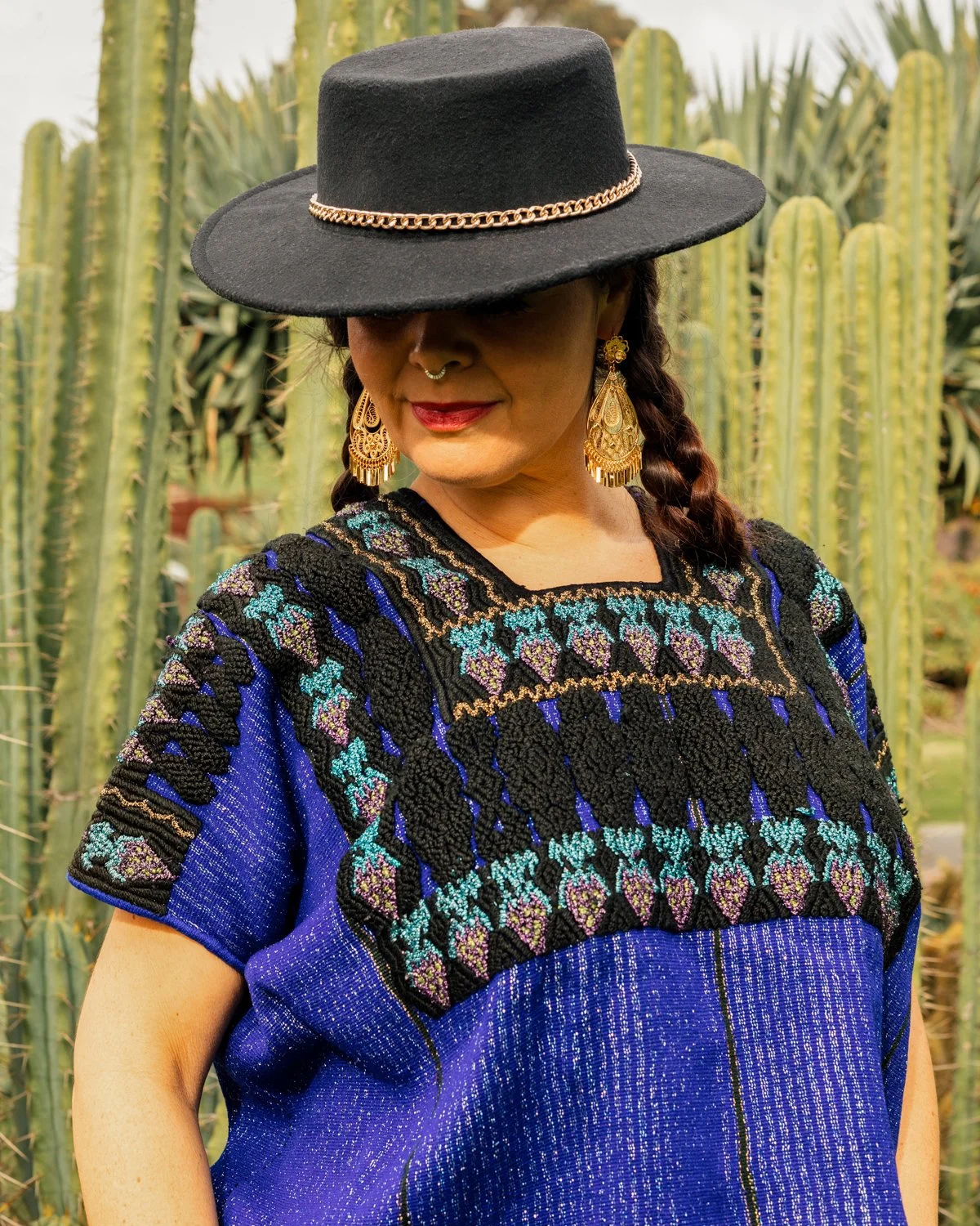 A woman wearing a wide-brimmed black hat with a gold chain, gold earrings, and a colorful embroidered blue dress, standing in front of tall cactus plants.