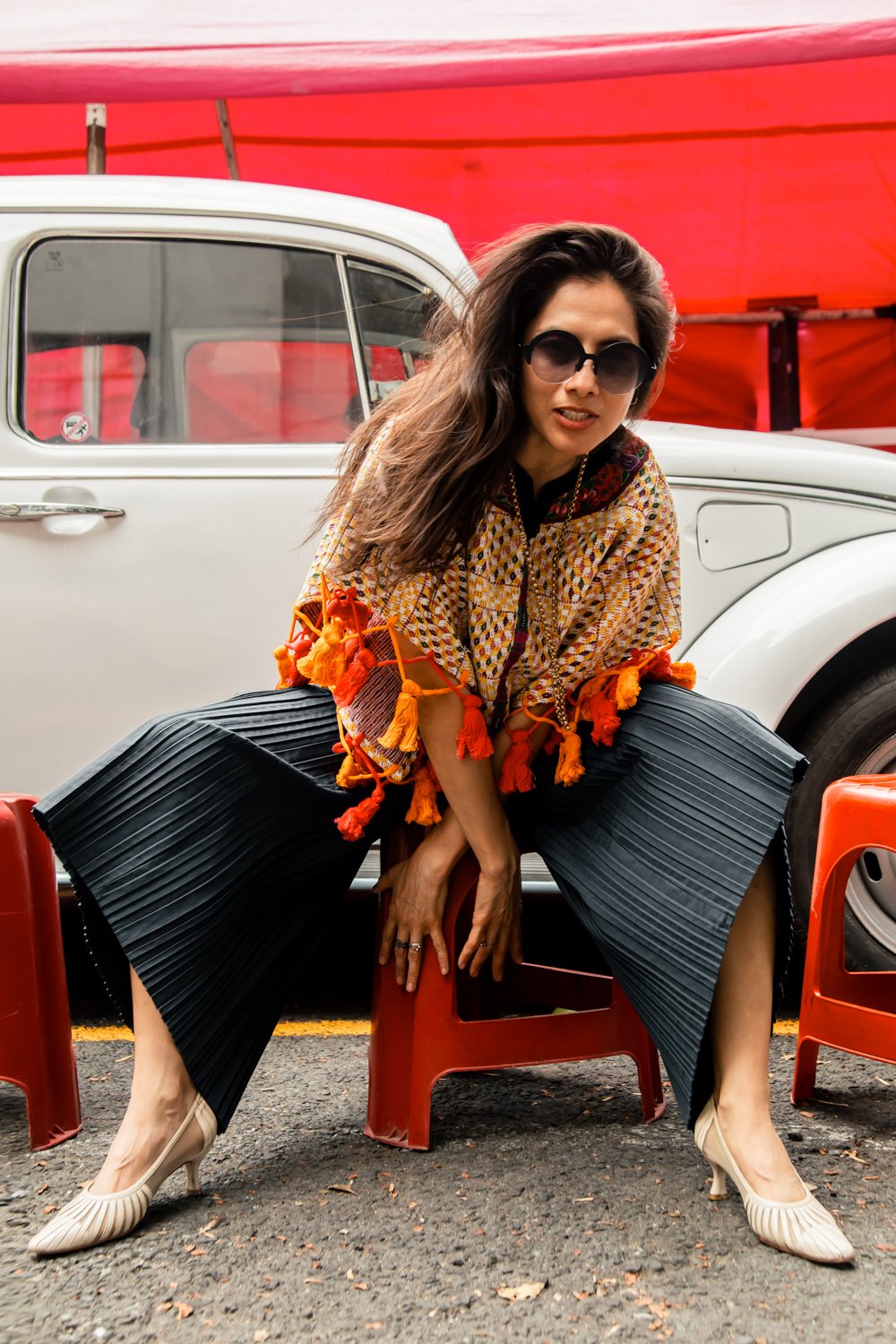 A woman with long dark hair, wearing black sunglasses, a patterned Mexican textile top with orange tassels, blue pleated wide-leg pants, and beige high heels, crouches on red plastic chairs in front of a vintage white car with a red canopy overhead