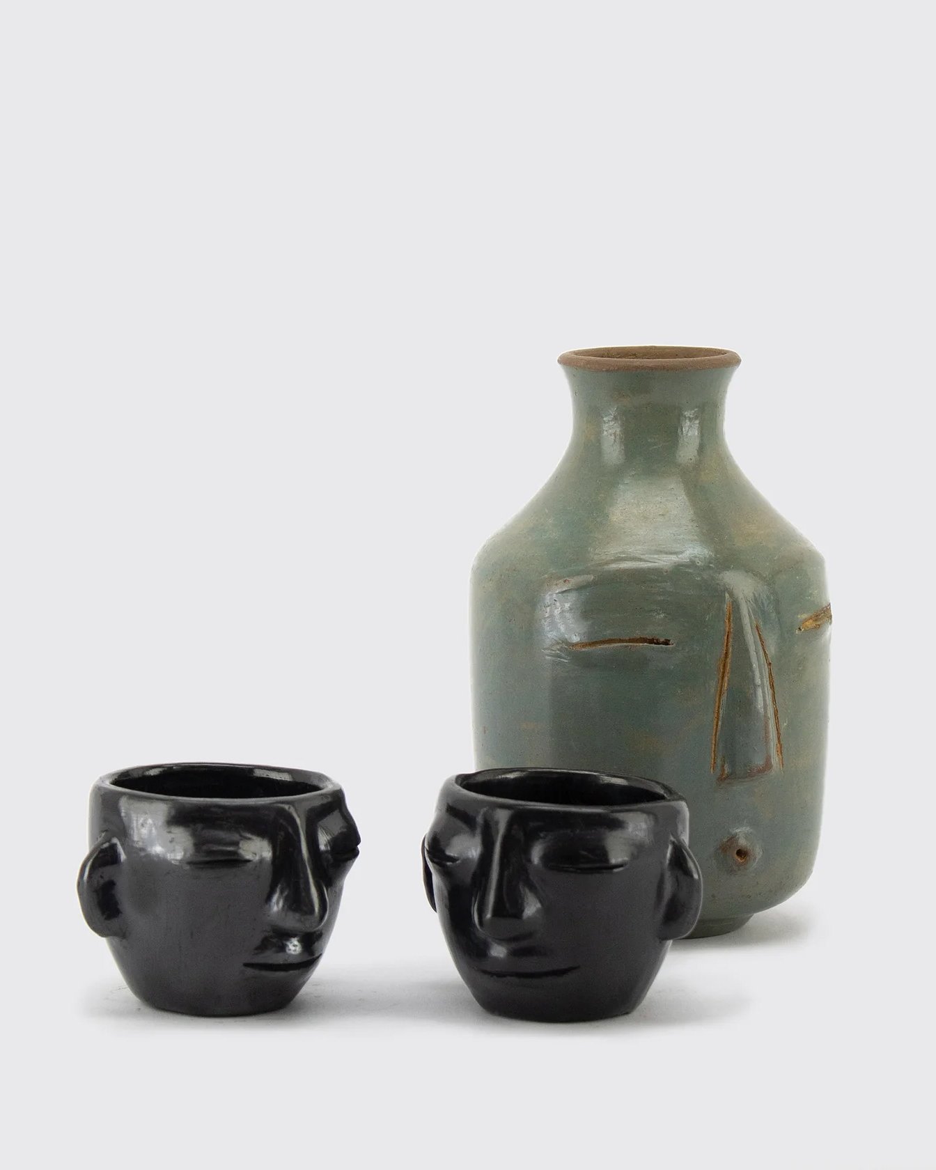 • A rustic handcrafted ceramic vase alongside traditional black burnished clay mezcal cups sculpted into human faces.