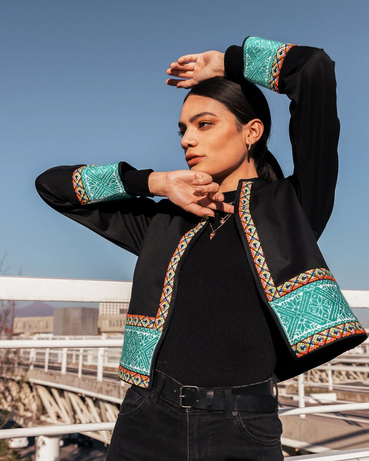 Young woman with dark hair wearing a black jacket with colorful embroidered accents, standing outdoors against a clear blue sky.