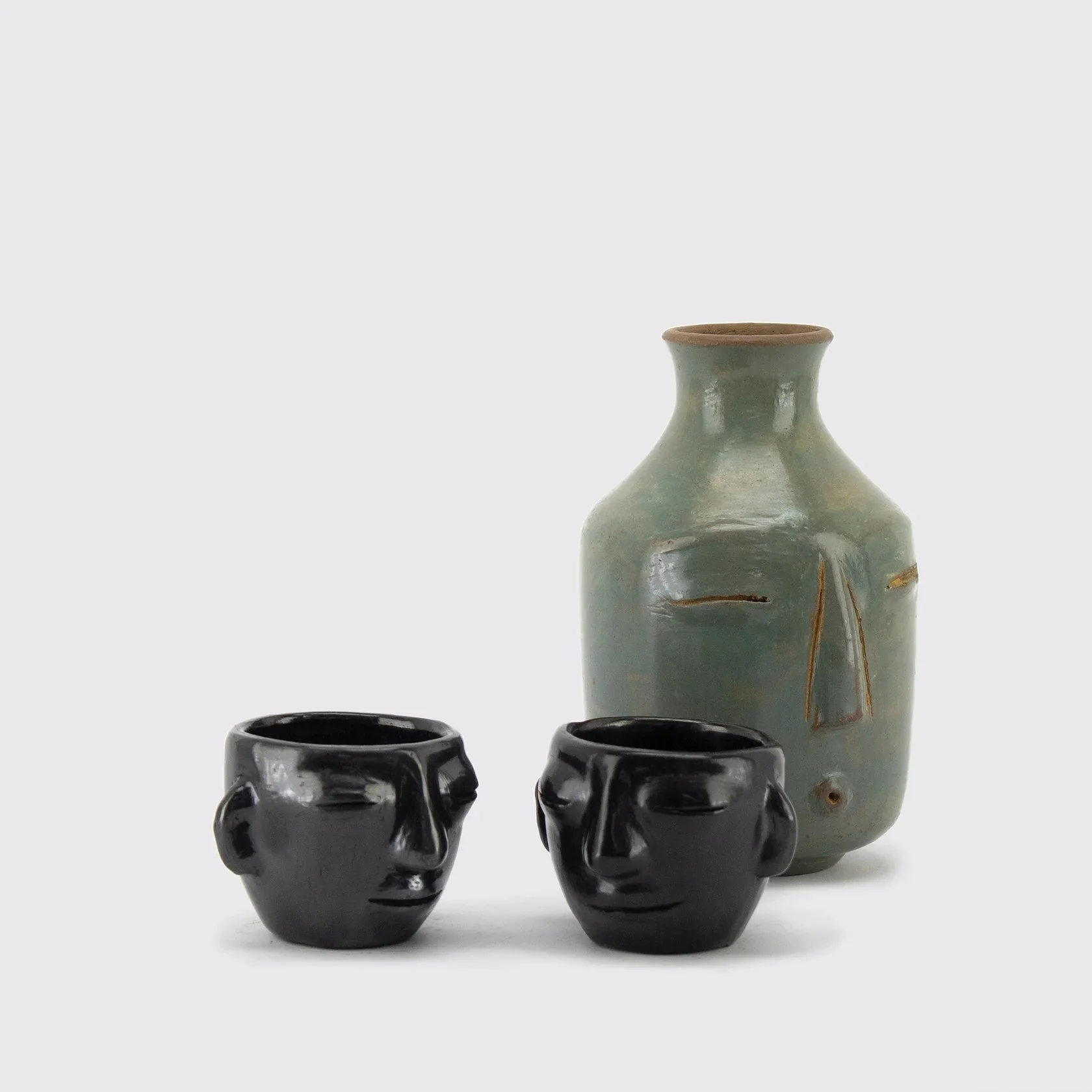 A ceramic bottle with a light green glaze and a narrow neck, flanked by three black skull-shaped cups.