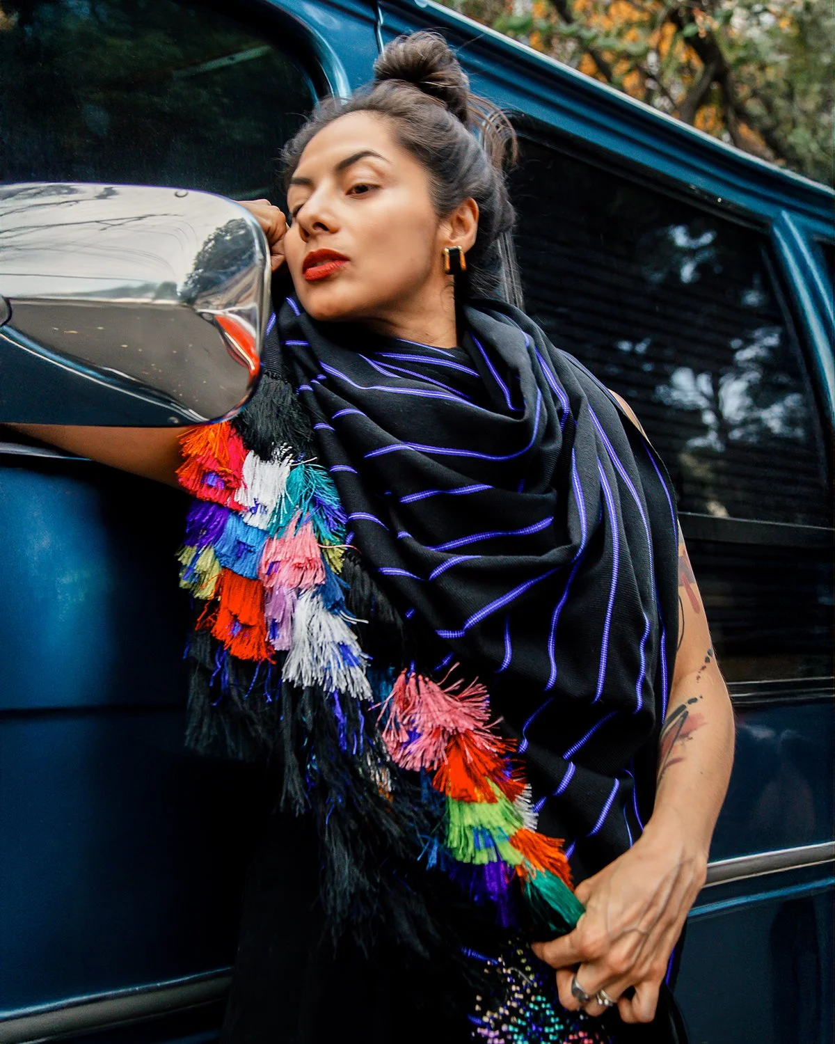 A woman with styled hair, makeup, and earrings leaning against a vehicle outdoors with trees in the background, wearing a colorful fringed scarf and a black dress.