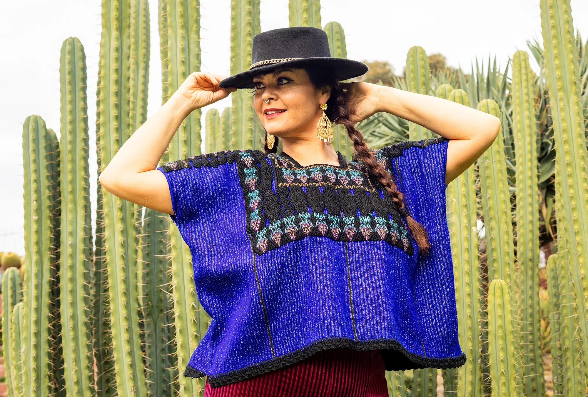 A smiling woman in a wide-brimmed black hat wearing a hand-embroidered blue Mexican huipil blouse in front of large green cacti.