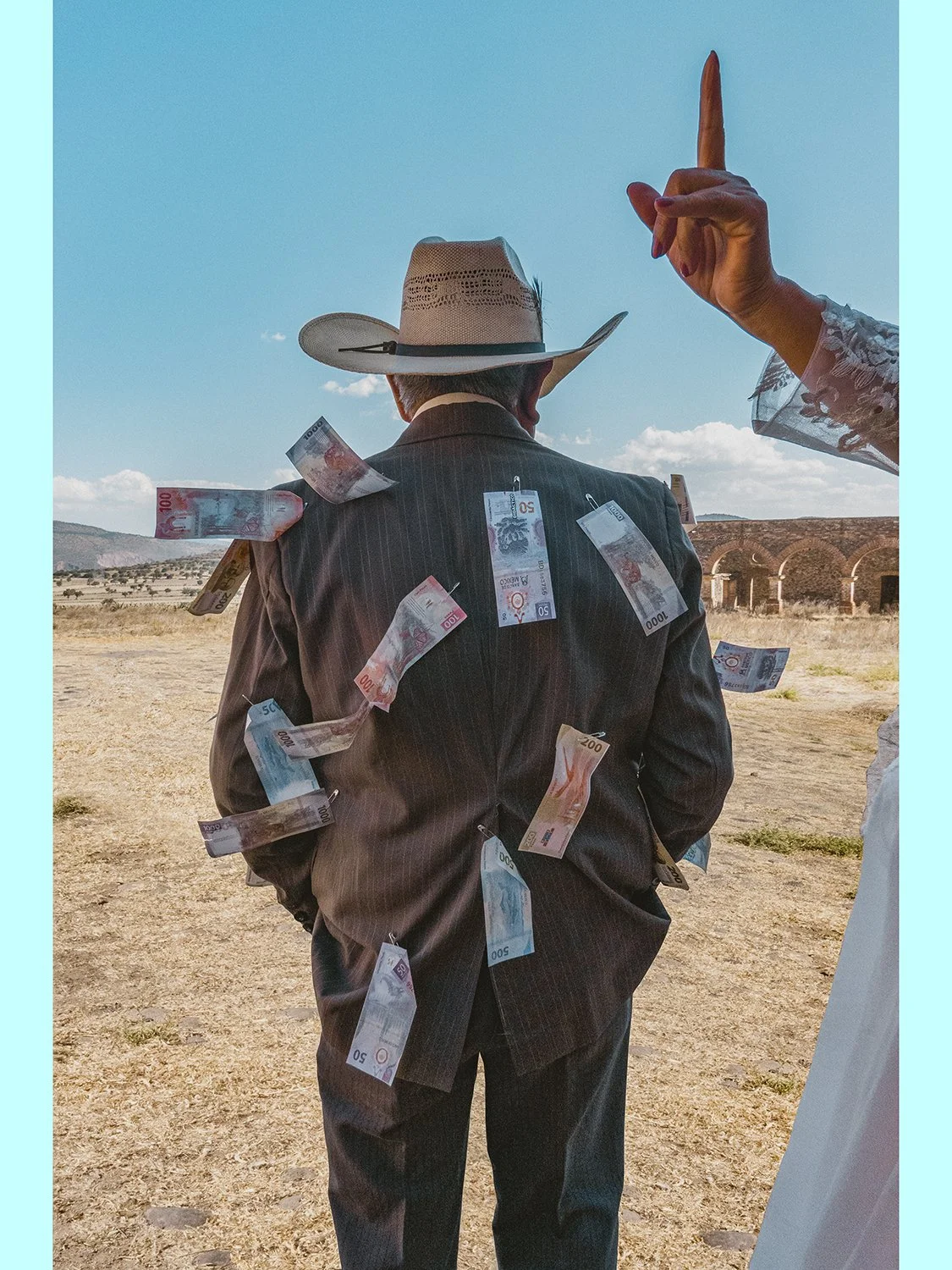Unframed fine art photography print of a man from behind wearing a cowboy hat and suit with Mexican banknotes pinned to his jacket during a wedding money dance.