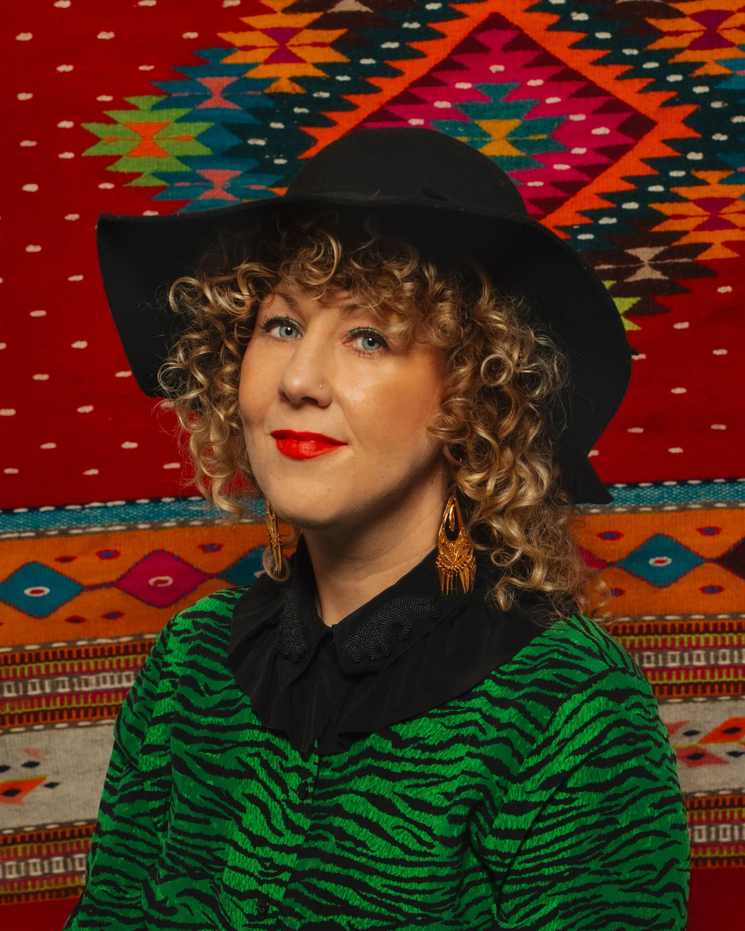 A woman with curly blonde hair and blue eyes wearing a wide-brimmed black hat, gold earrings, red lipstick, and a green and black animal print cardigan, standing in front of a colorful woven rug with geometric patterns.