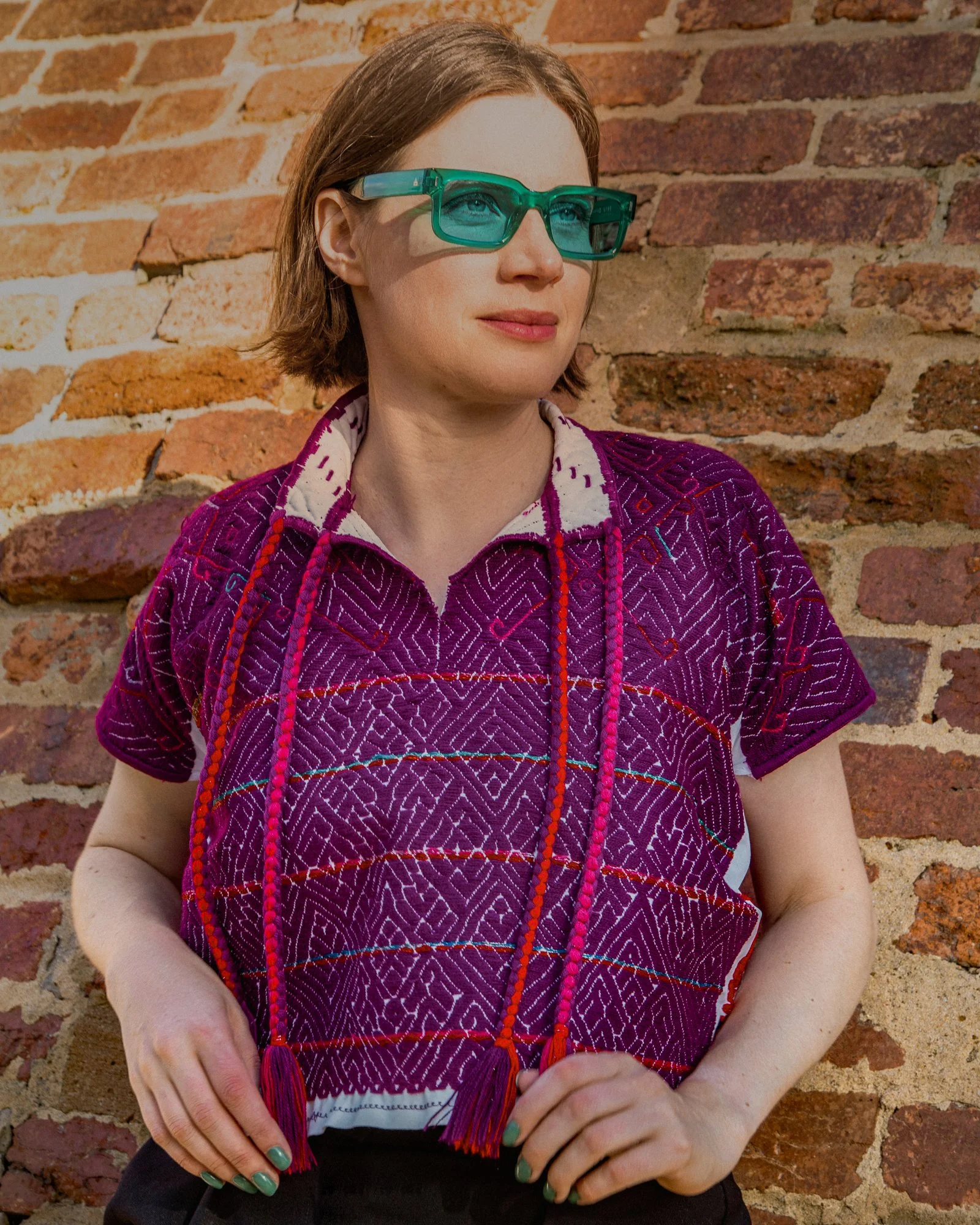 Woman with short brown hair wearing oversized teal glasses and a traditional embroidered purple garment, sitting against a brick wall.