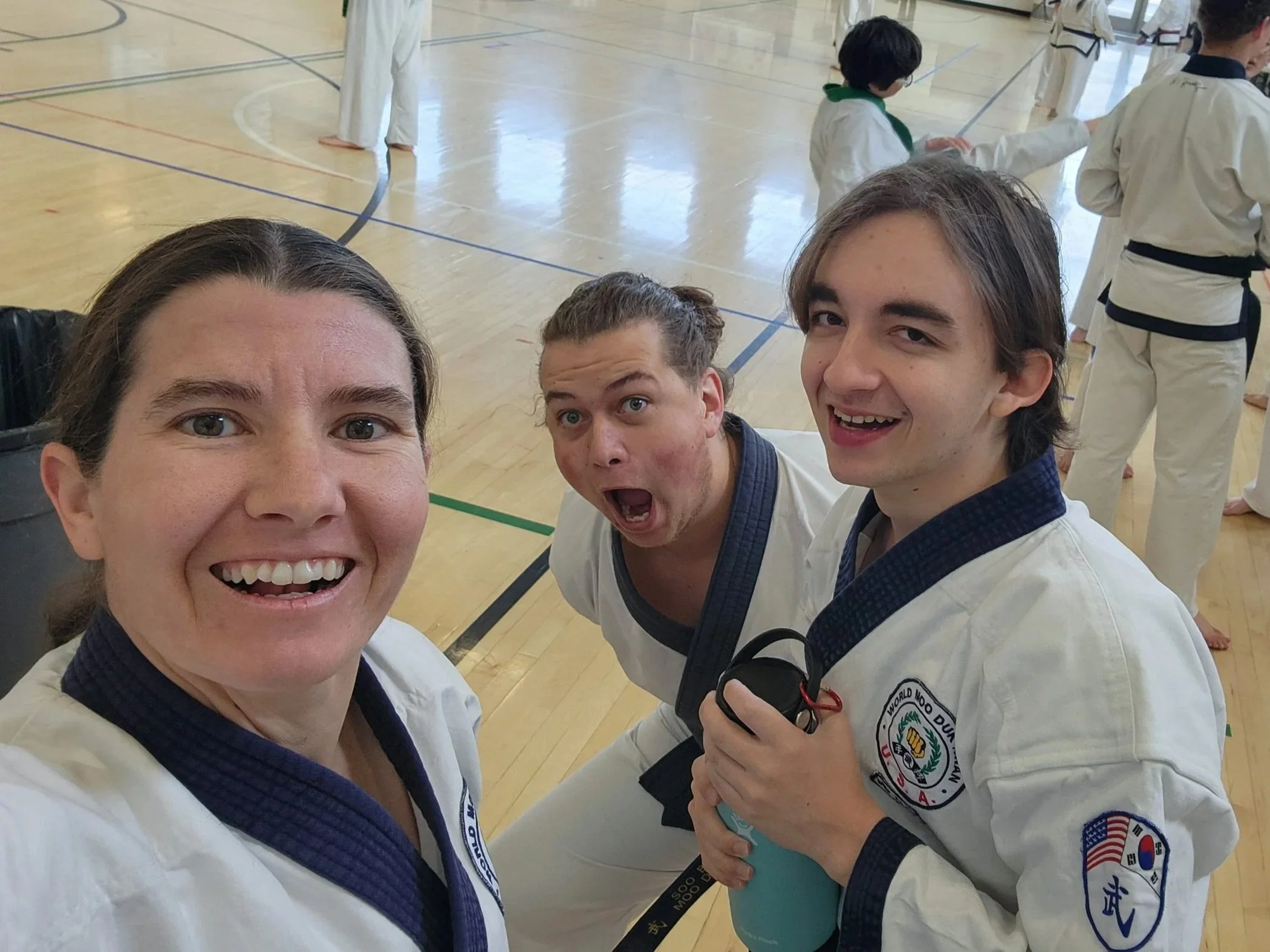 Selfie of four people in martial arts uniforms at a gym, with additional people practicing martial arts in the background.