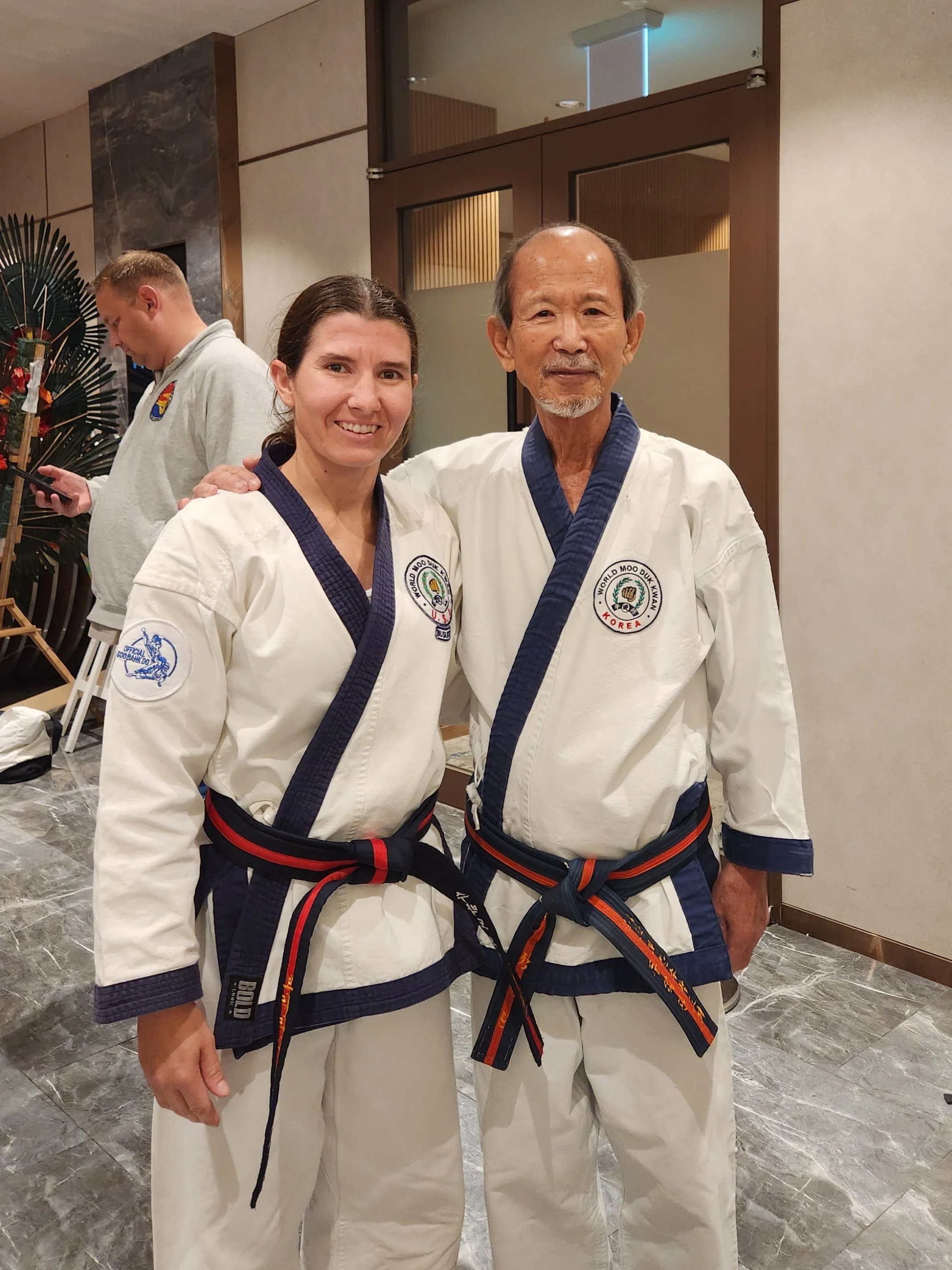Two martial artists in traditional uniforms, smiling and standing side by side, in a room with a marble floor and a decorative wall.