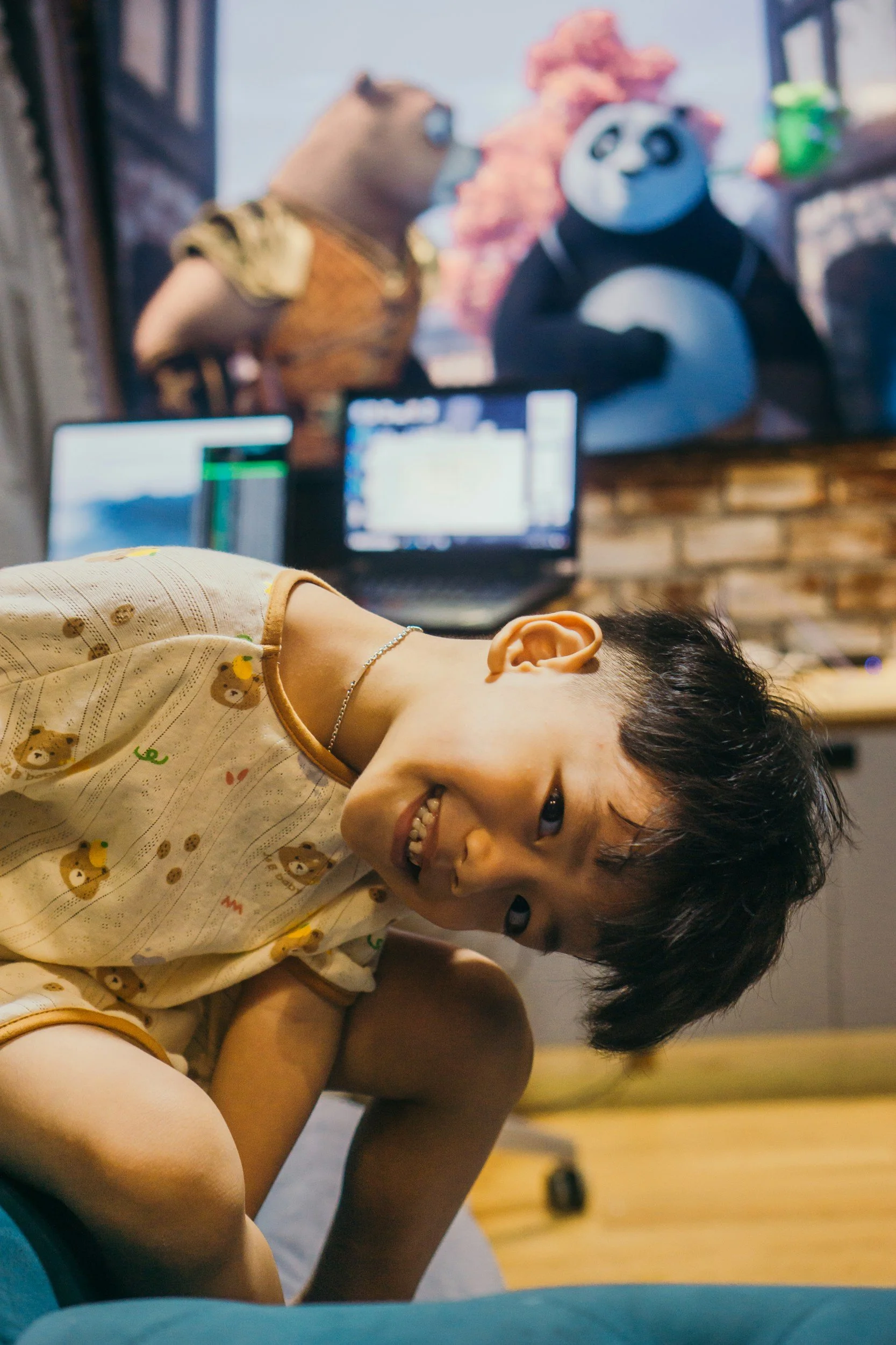 Stock image of a young boy smiling and lying on the floor in a game or activity area, with a large screen and colorful puppets of an owl, panda, and a bear in the background.