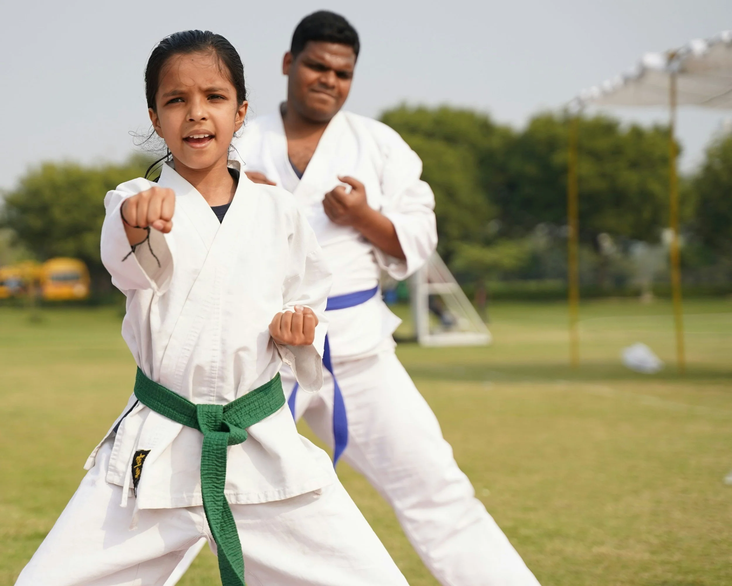 Stock photo of two children practicing martial arts outdoors, wearing traditional white martial arts uniforms with colored belts, one with a green belt and the other with a blue belt, in a park with trees and a sports field in the background.