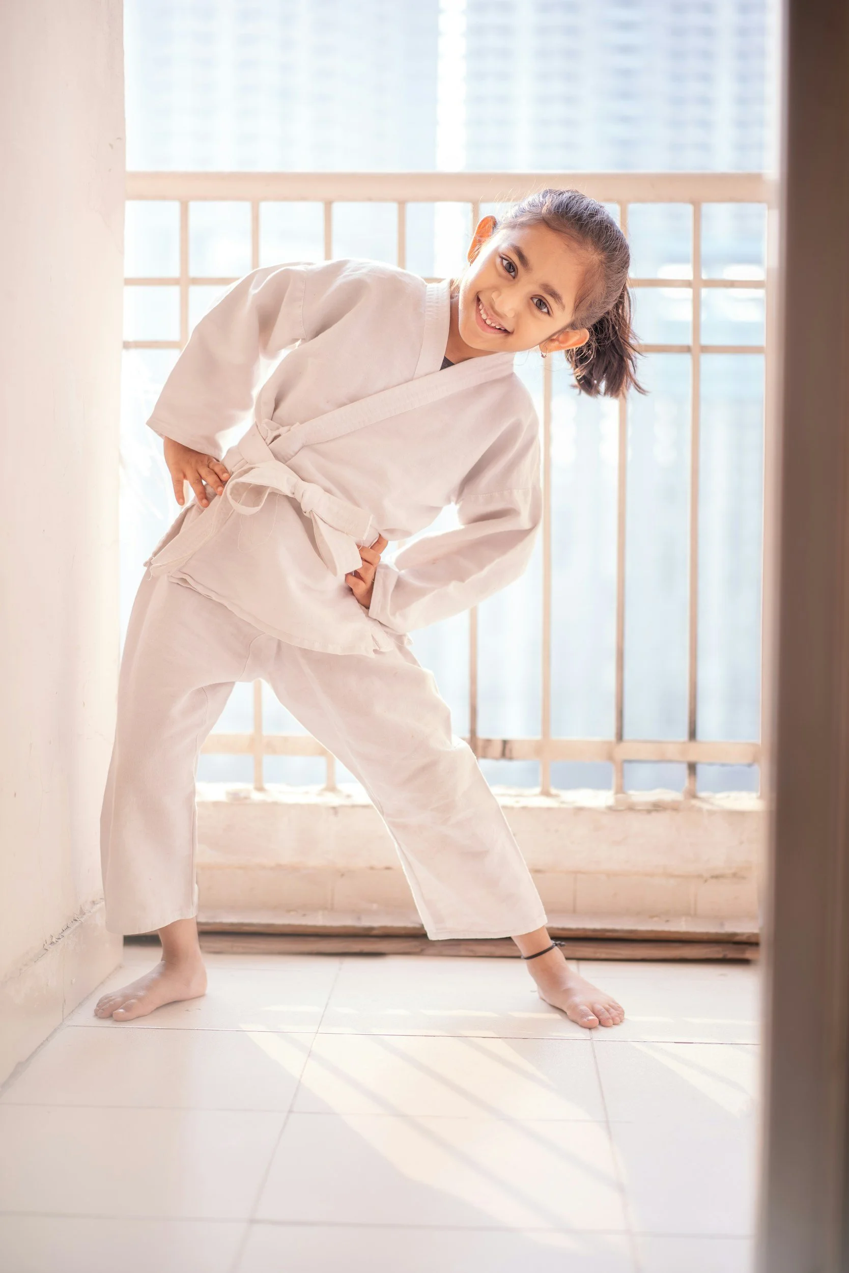 Stock image of a young girl practicing martial arts or yoga in a bright room, wearing a white gi, striking a pose with hands on hips and smiling at the camera.