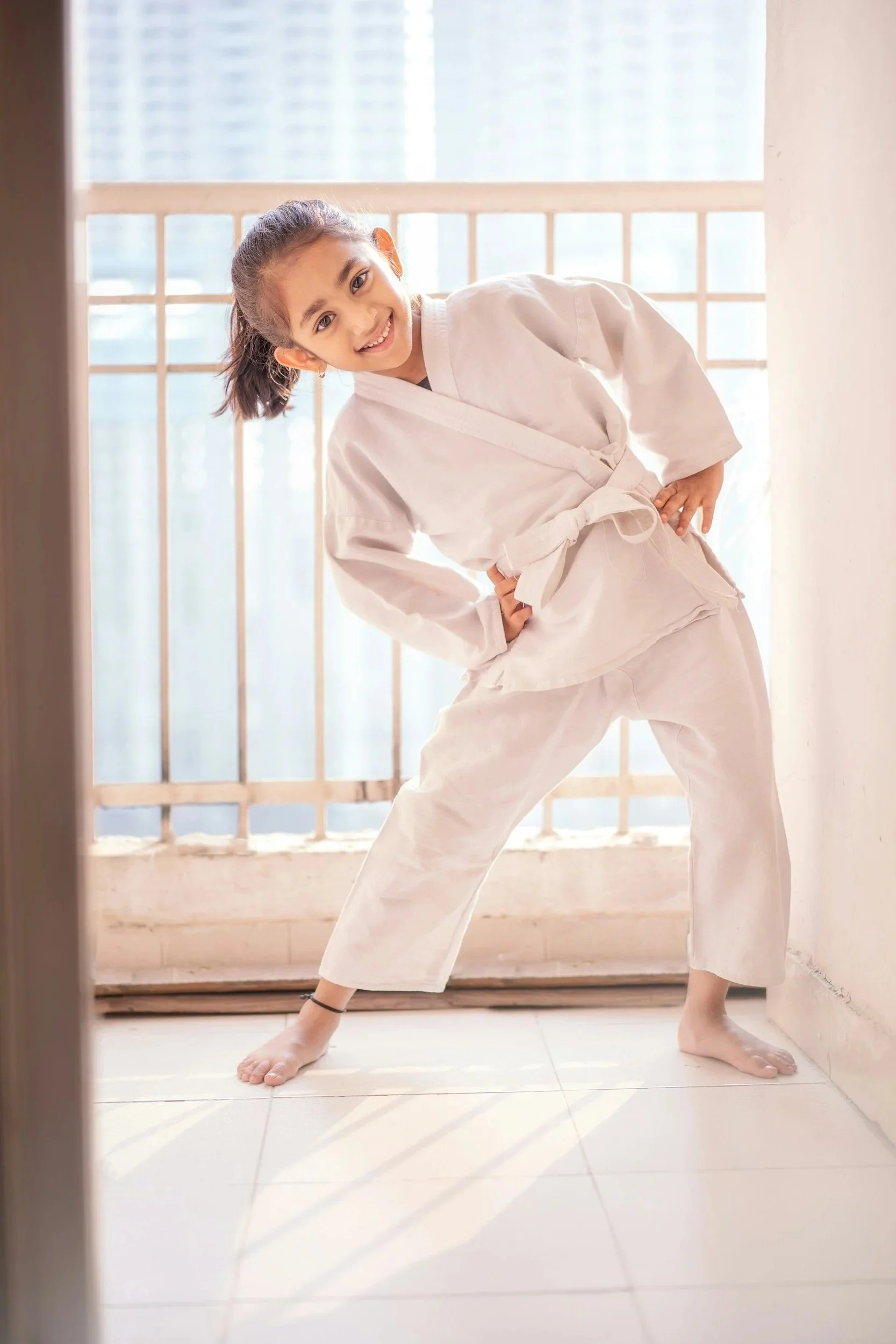 Stock image of a young girl in a white martial arts gi practicing martial arts indoors near a window, smiling and posing confidently.