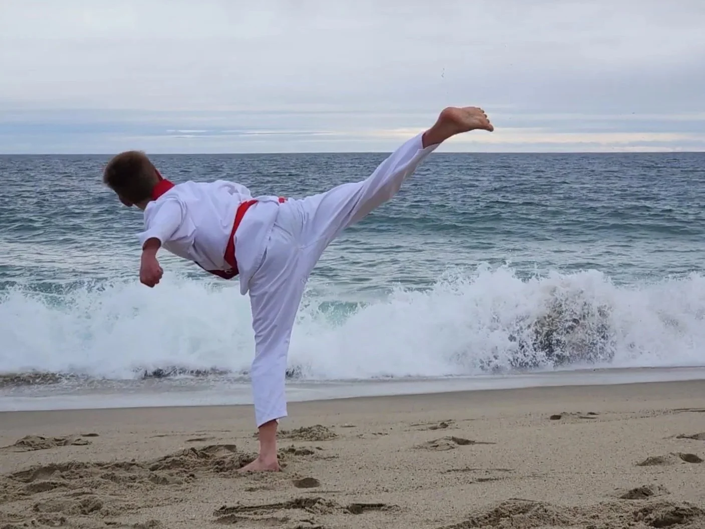 A person practicing martial arts on a beach, performing a high kick near the water.