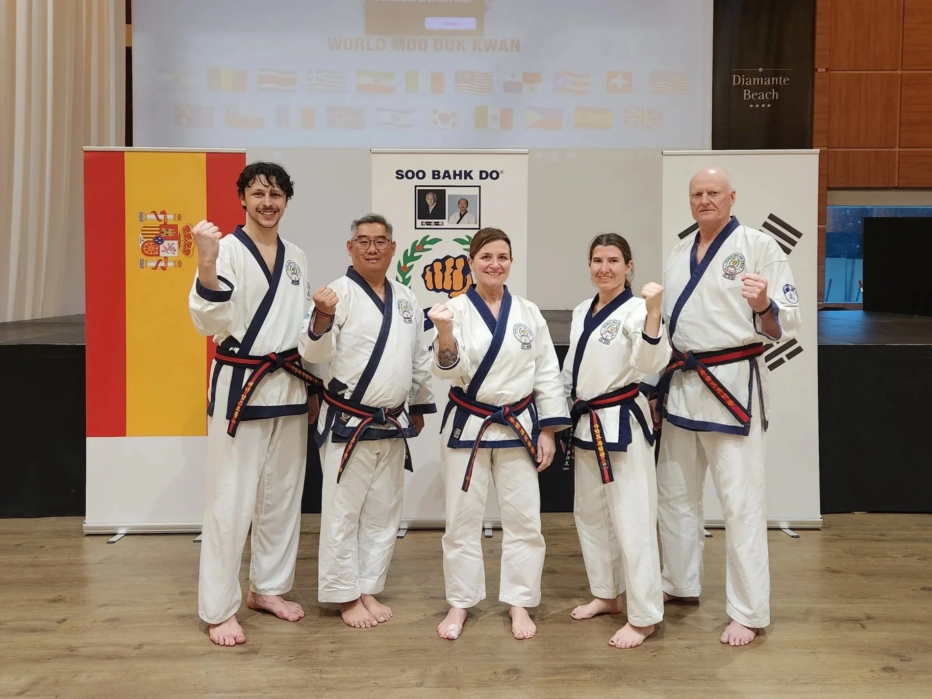 Group of five martial artists in white uniforms with black belts, standing indoors on a wooden floor, posing with fists raised. Behind them, flags from Spain and South Korea are visible, along with banners and a large screen displaying flags from multiple countries.