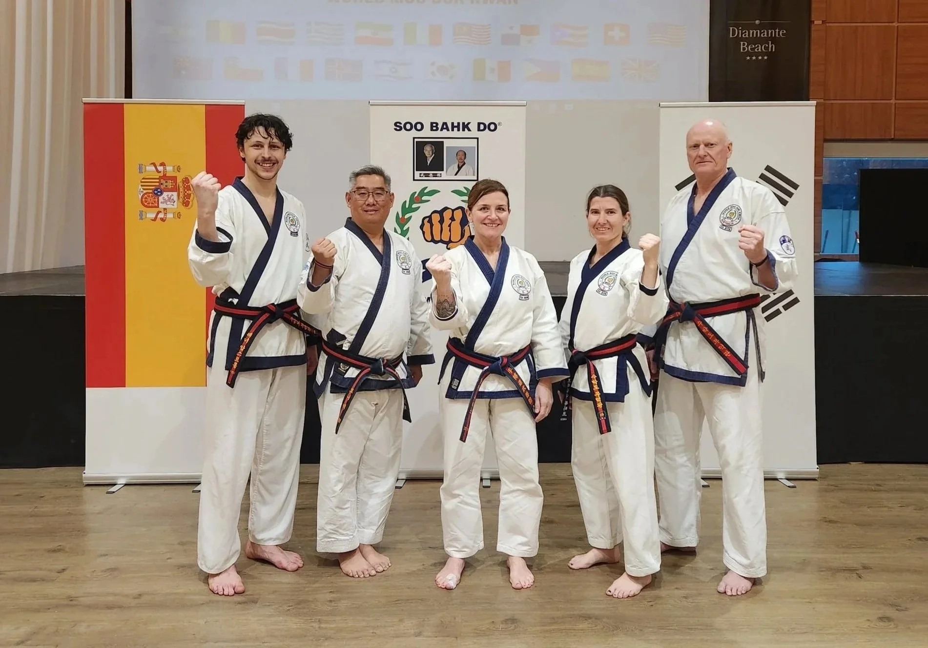 Five martial artists in white uniforms with black and red belts standing on a wooden floor, posing with fists clenched and smiling. Banners and flags from Spain, Korea, and other countries are in the background, along with a screen above displaying various flags.