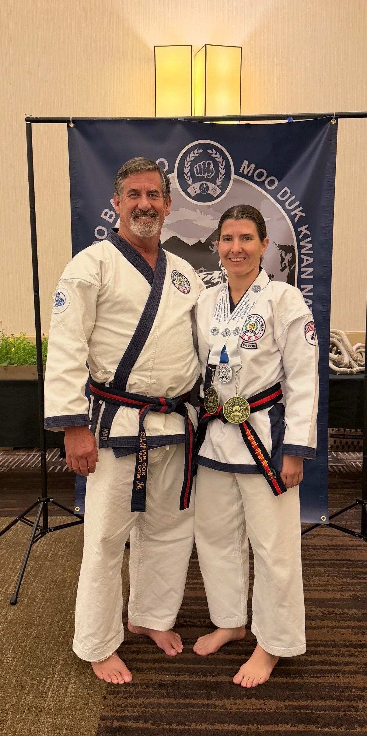 Two martial artists in white uniforms with blue and red ko dan ja belts standing together with medals, in front of a martial arts banner.