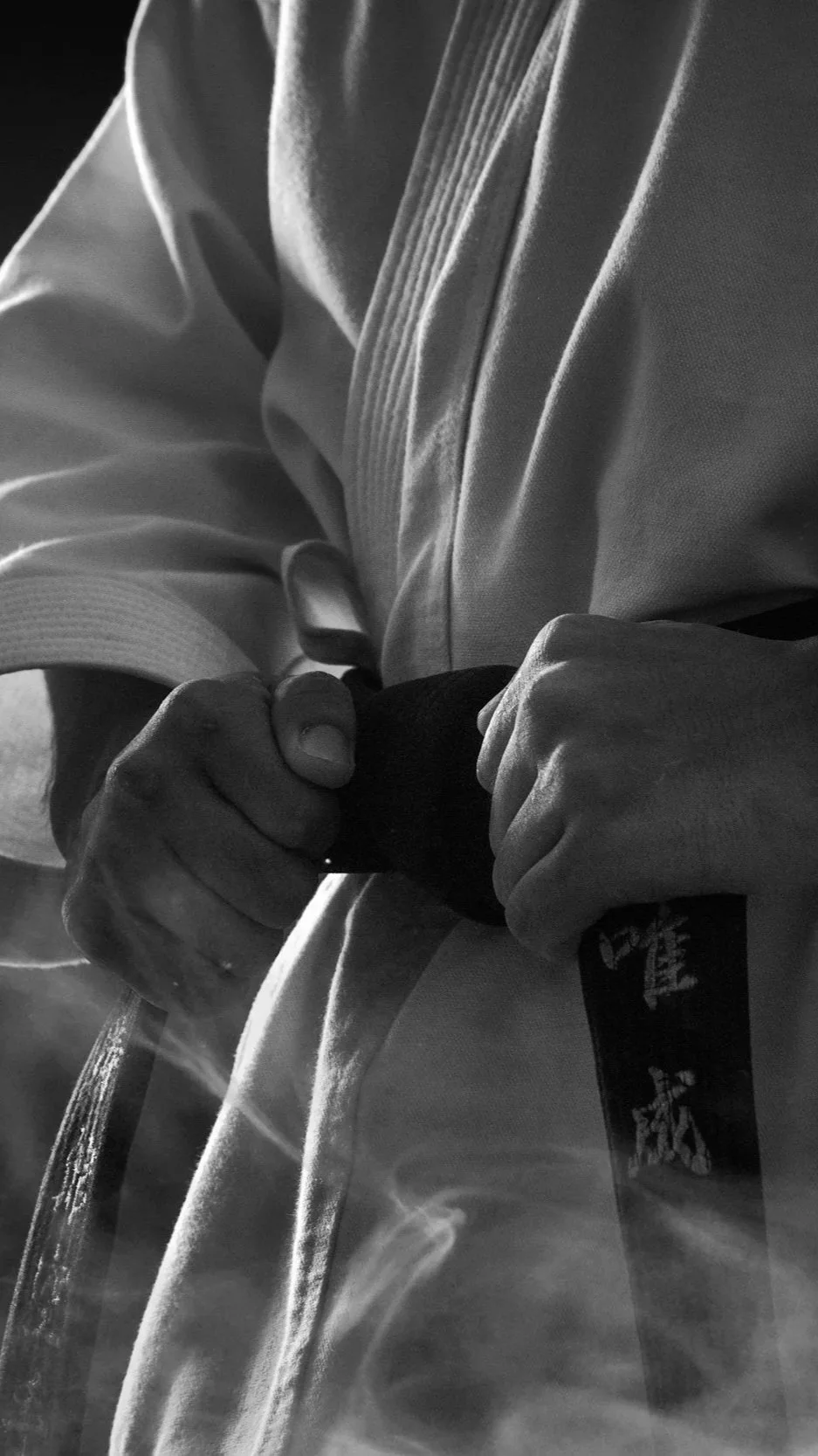 Stock image of a person dressed in a martial arts uniform, or gi, tying a black belt around their waist.