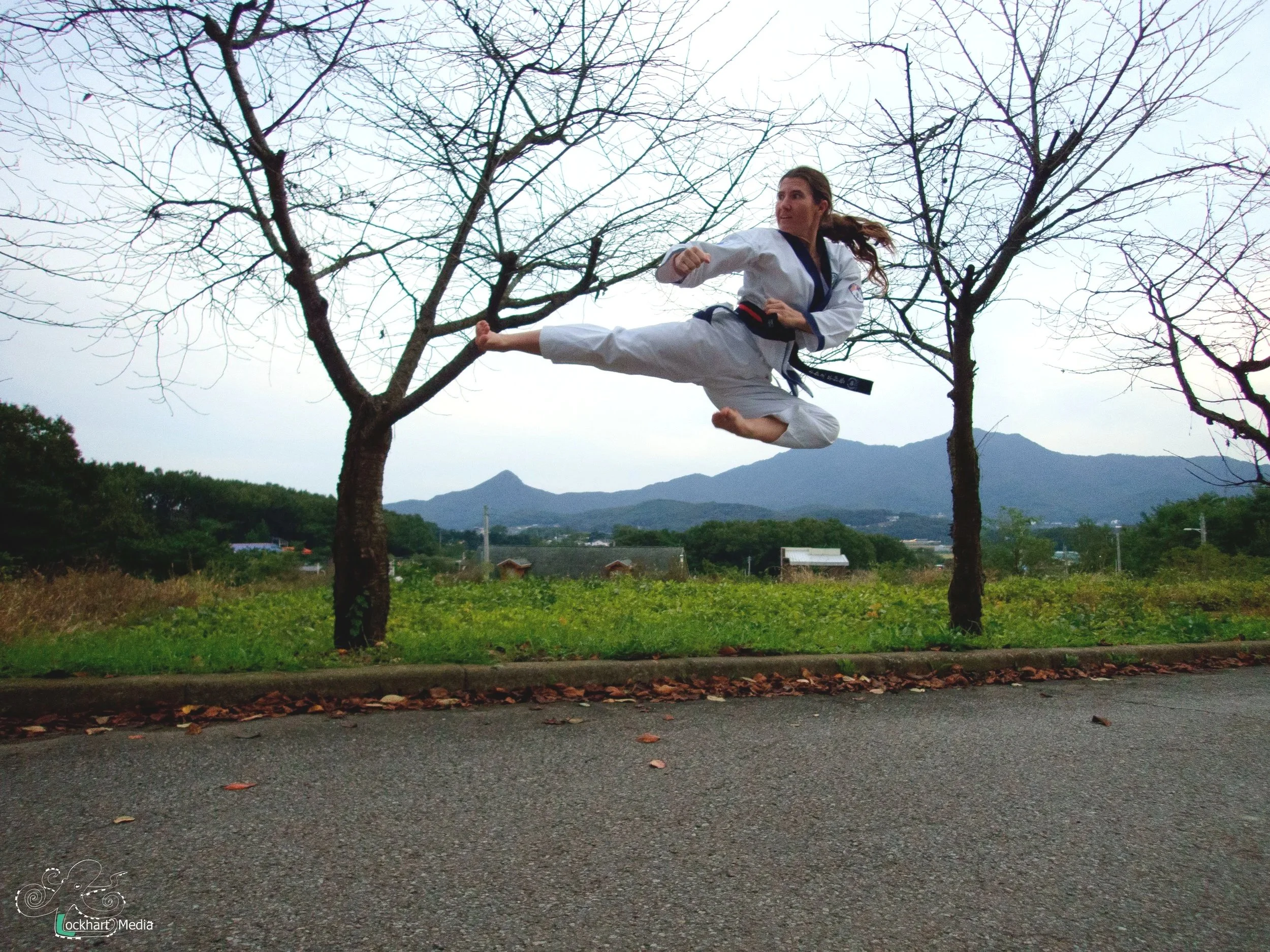 A woman dressed in a martial arts uniform is performing a flying kick outdoors. She is in mid-air between two leafless trees with mountain peaks and a cloudy sky in the background. The ground below is paved with a strip of leaves along the edge.