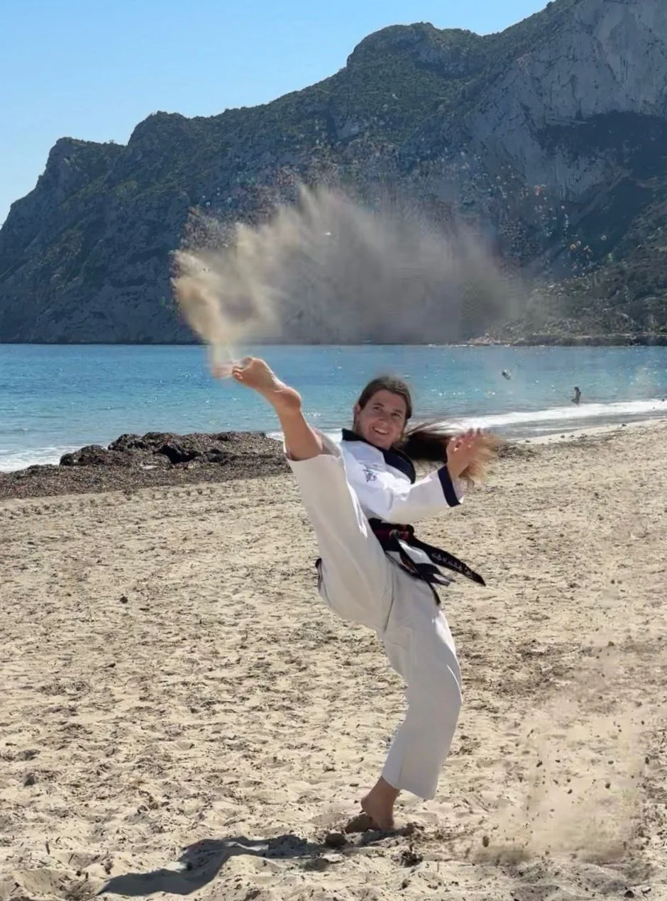 Person in a martial arts uniform practicing a kick on a sandy beach with mountains, water, and a person in the background.
