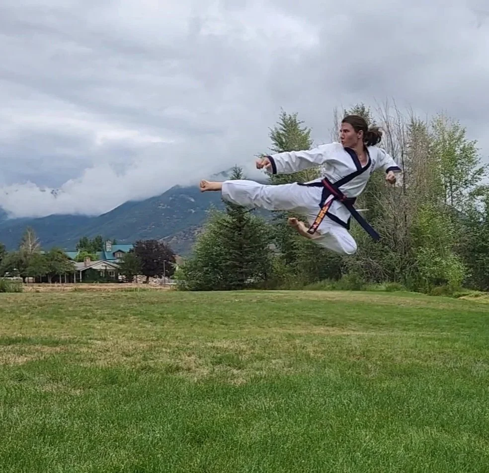 A woman performing a high side kick in a martial arts uniform outdoors during daytime, with mountains and trees in the background.