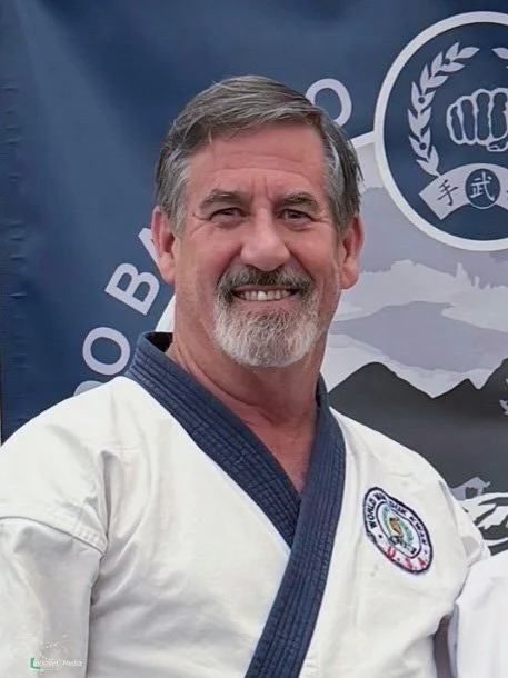 A smiling man in a martial arts gi stands in front of a martial arts banner.
