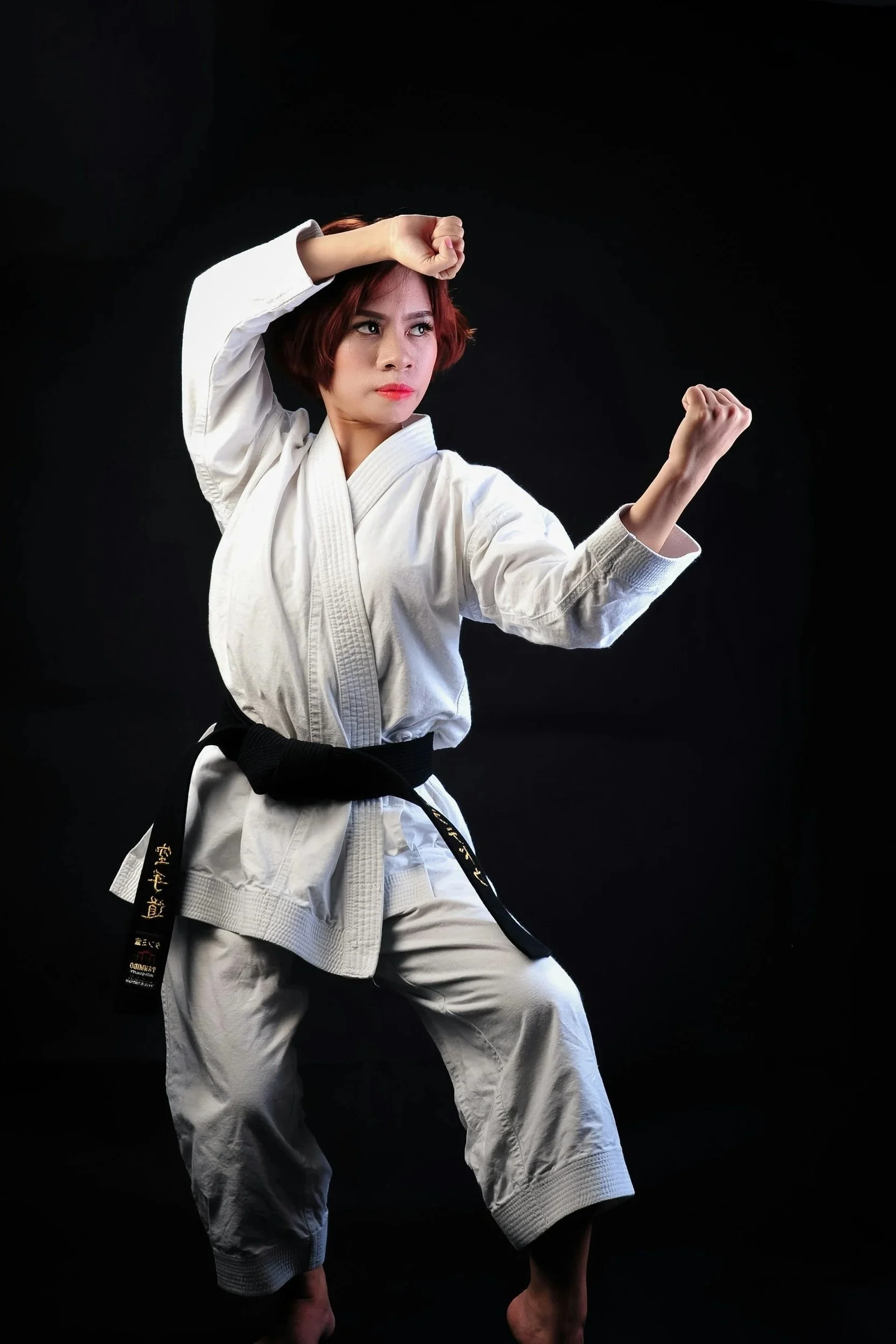 Stock image of a woman dressed in a white martial arts gi with a black belt, performing a martial arts stance against a black background.