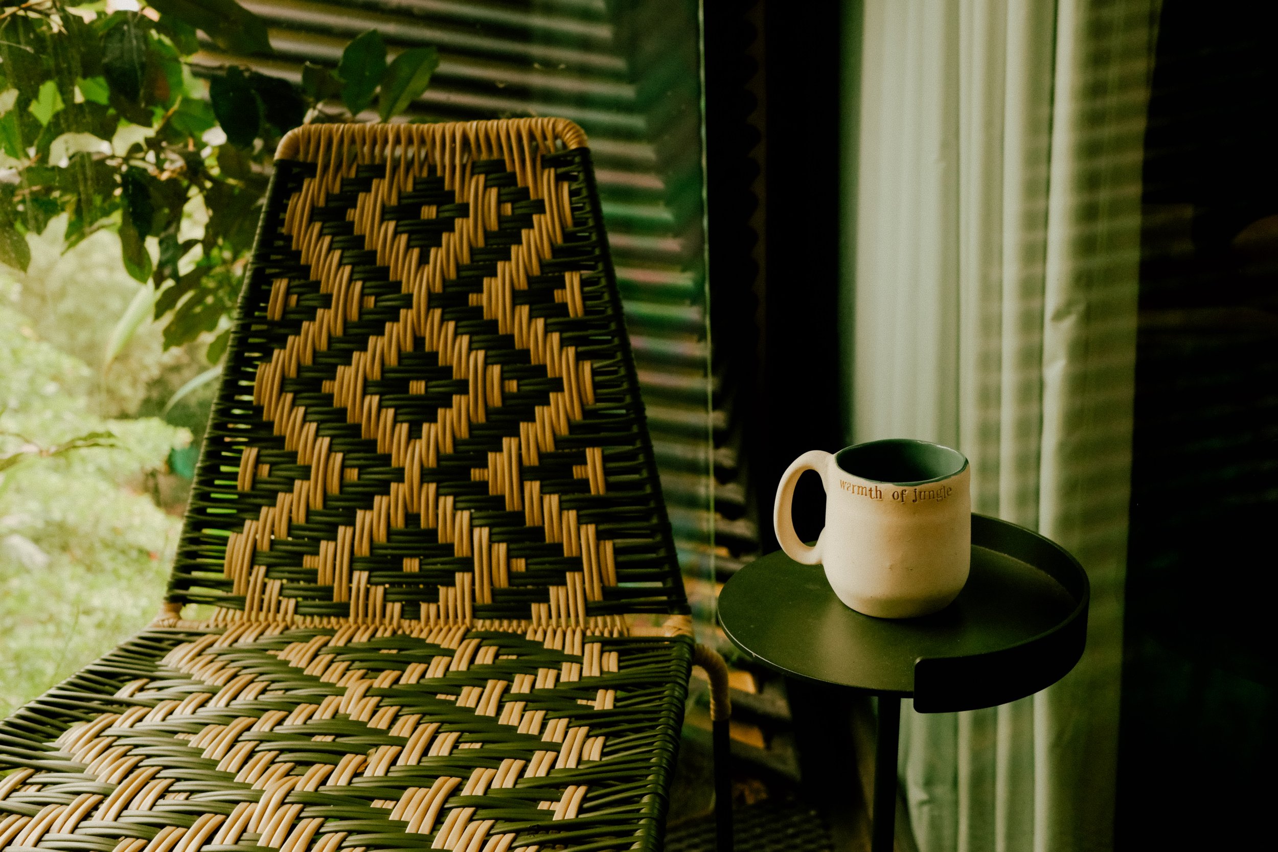 A wicker chair with a black and beige diamond pattern on the backrest, placed next to a black round table with a white mug that has text reading 'warmth of jungle', set against a window with green blinds and sheer curtains.