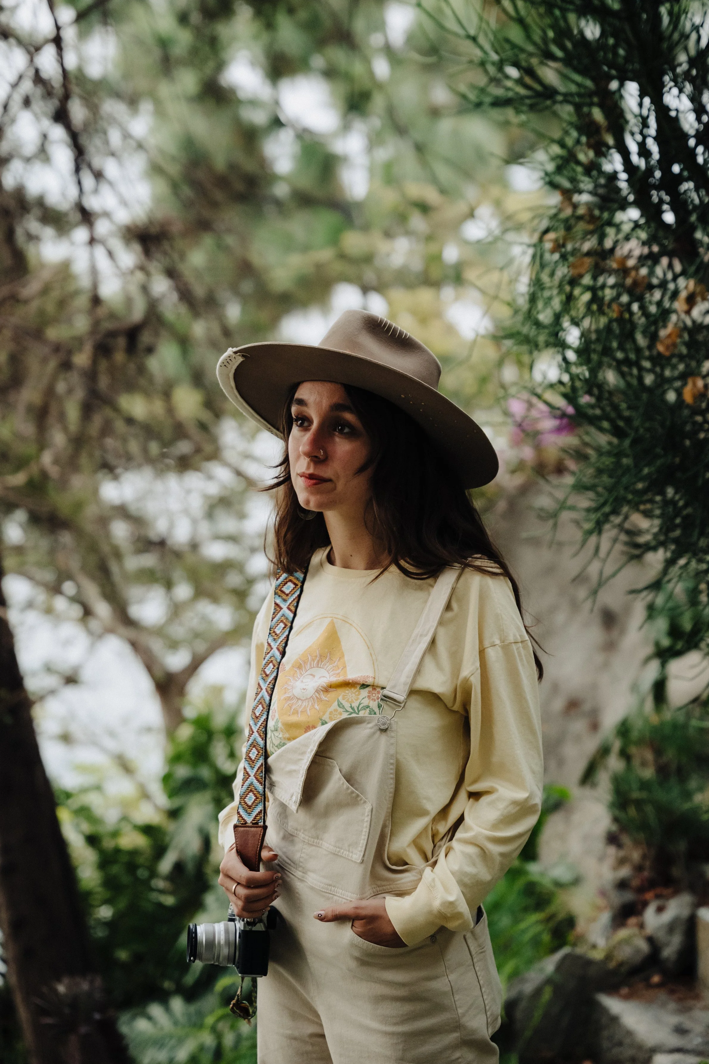 A woman wearing a wide-brimmed hat and a beige jacket stands outdoors with greenery in the background, holding a camera.