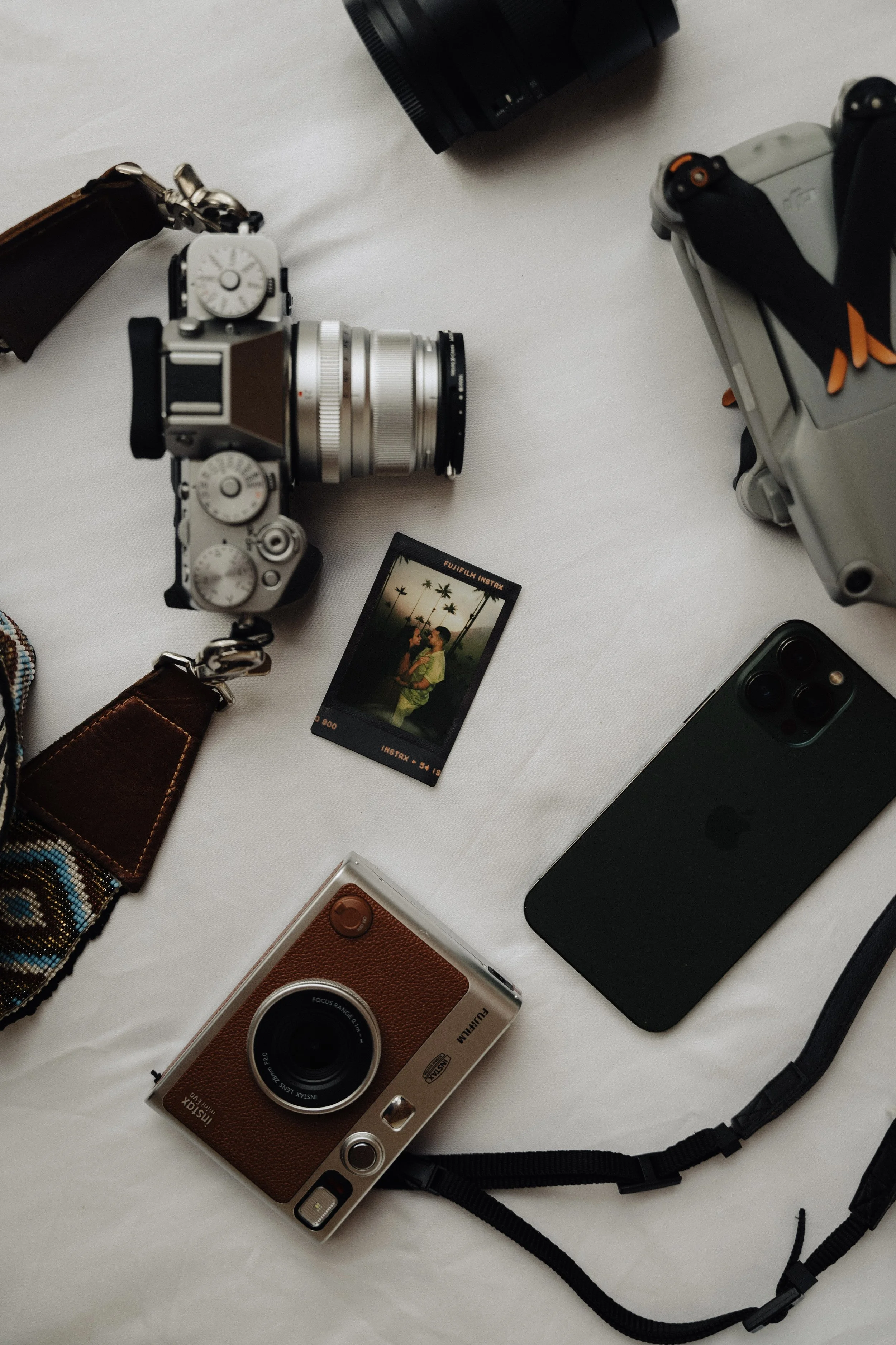 A flat lay of various photography and tech equipment on a white surface, including a vintage film camera, a modern digital camera, a smartphone, a camera lens, a photo print, a leather camera strap, and a protective case.