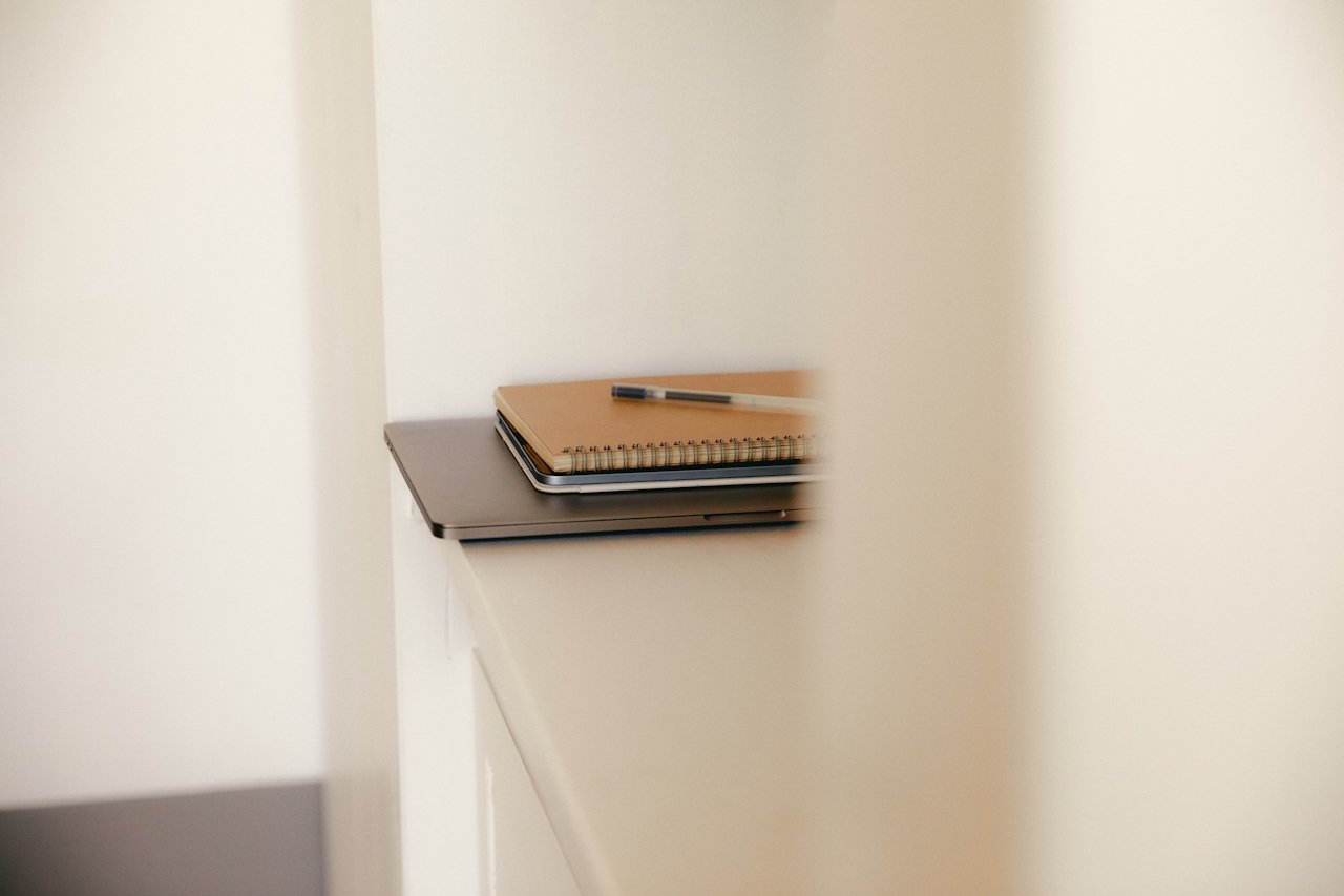 A closed notebook with a pen on top, placed on a desk or shelf near a white wall.